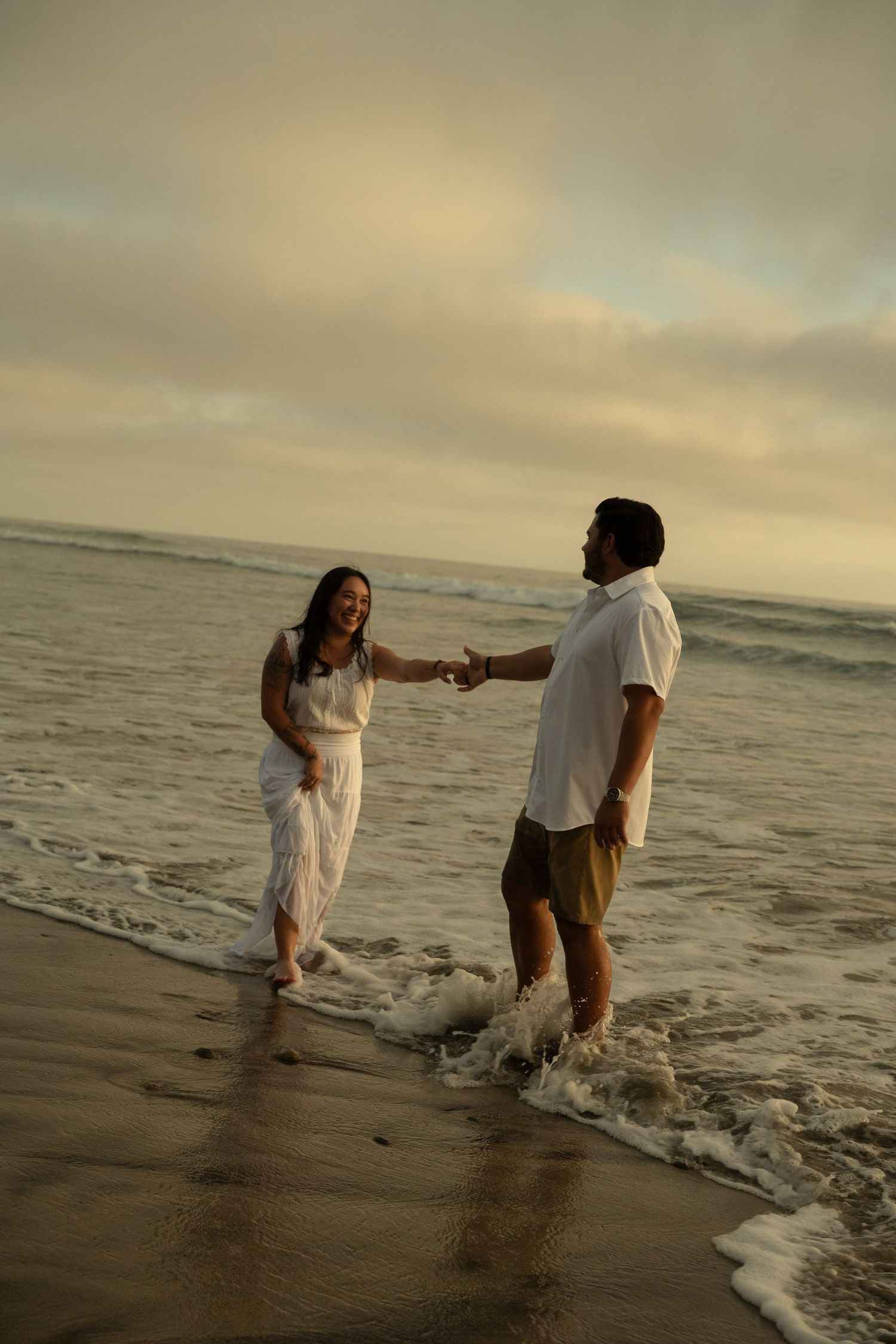 Joyful couple holding hands and splashing in ocean waves at sunset, woman in white dress laughing on sandy beach.
