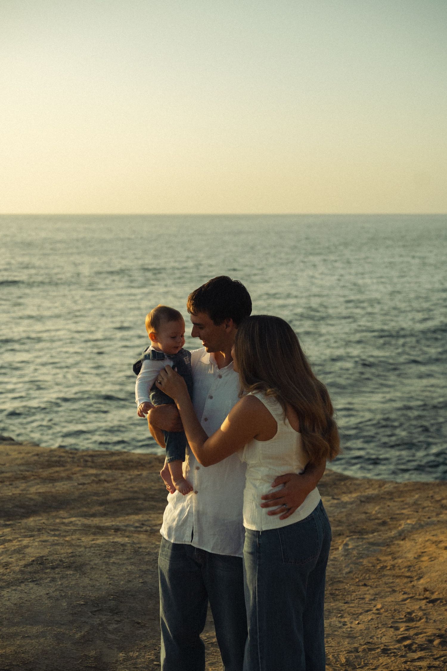Young family of three sharing a tender moment together on a rocky beach at sunset.