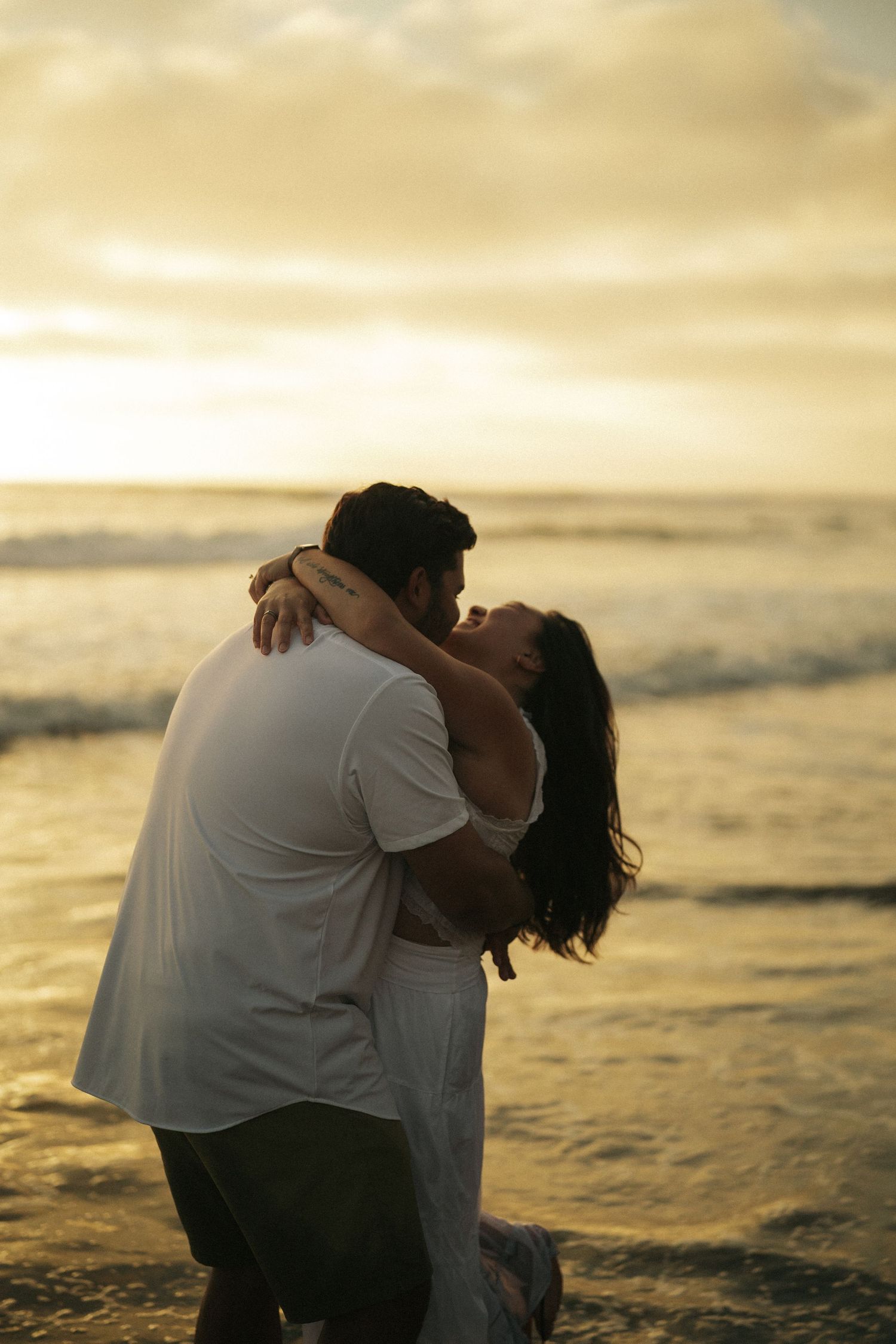 A couple embraces romantically on a beach at golden sunset, waves crashing behind them as woman laughs joyfully.
