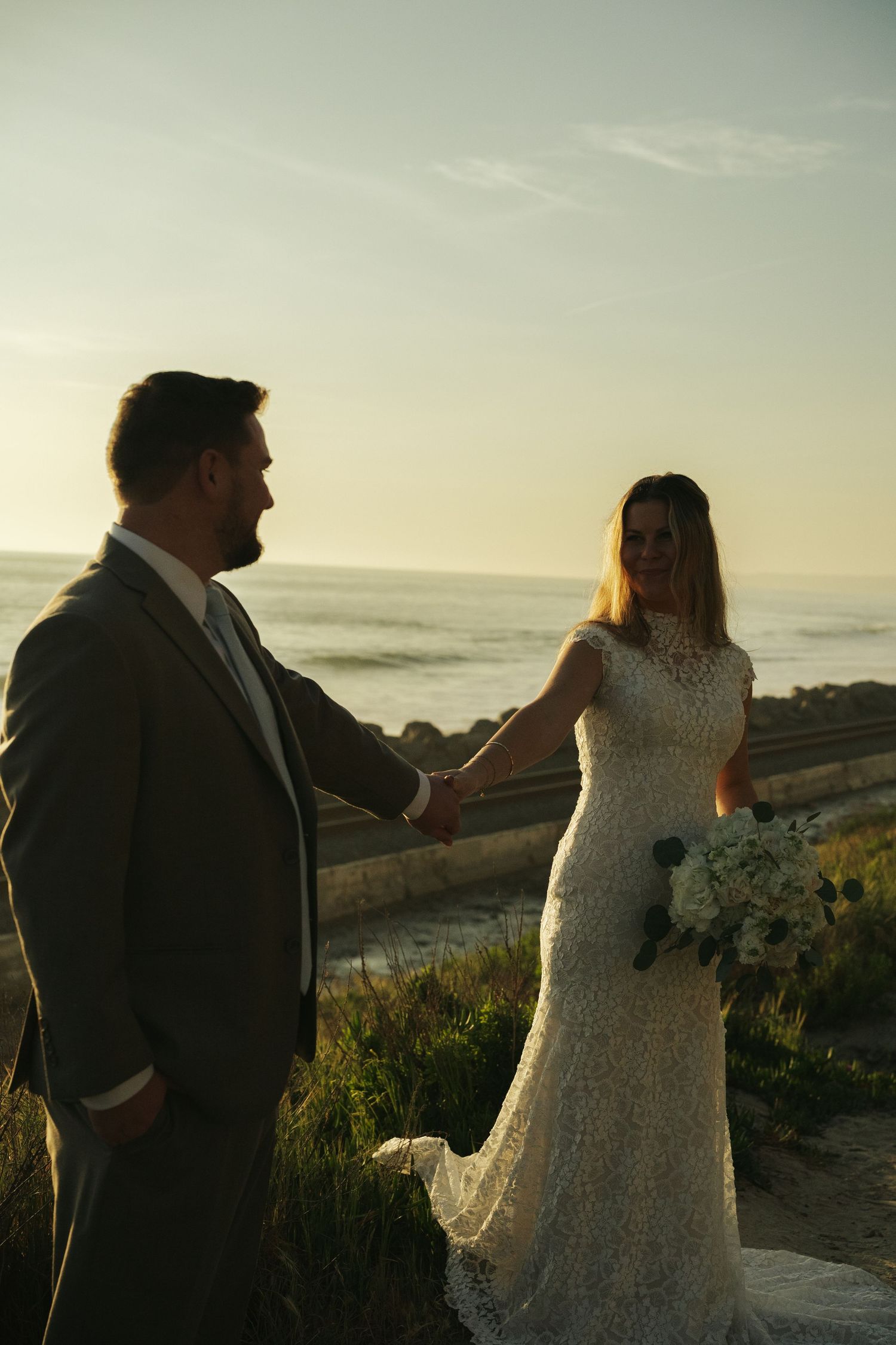 Bride and groom hold hands at sunset near rocky ocean shore, bride wearing lace gown holding white bouquet.