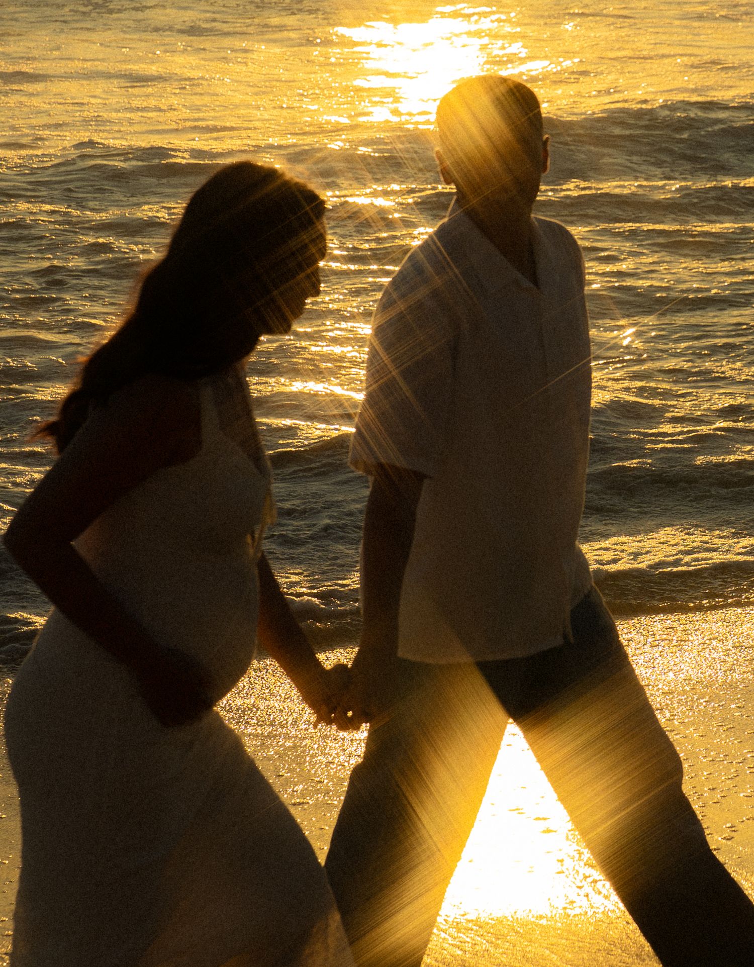 A couple in wedding attire holds hands walking on a beach at golden sunset, silhouetted against shimmering ocean waves.