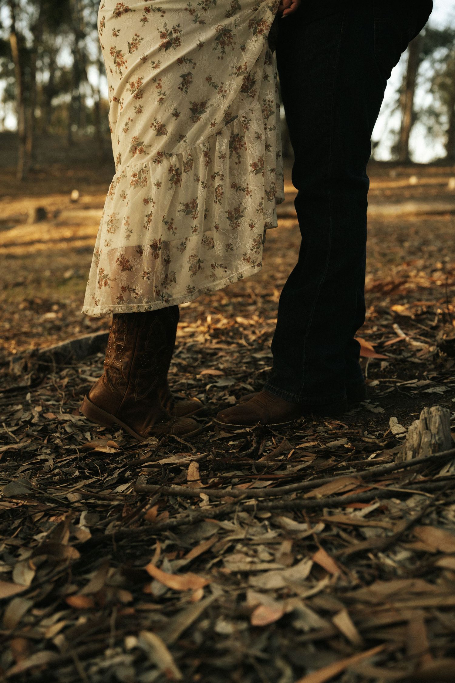 Couple standing close together in autumn forest, woman in floral dress and brown boots, man in dark jeans on leaf-covered ground.