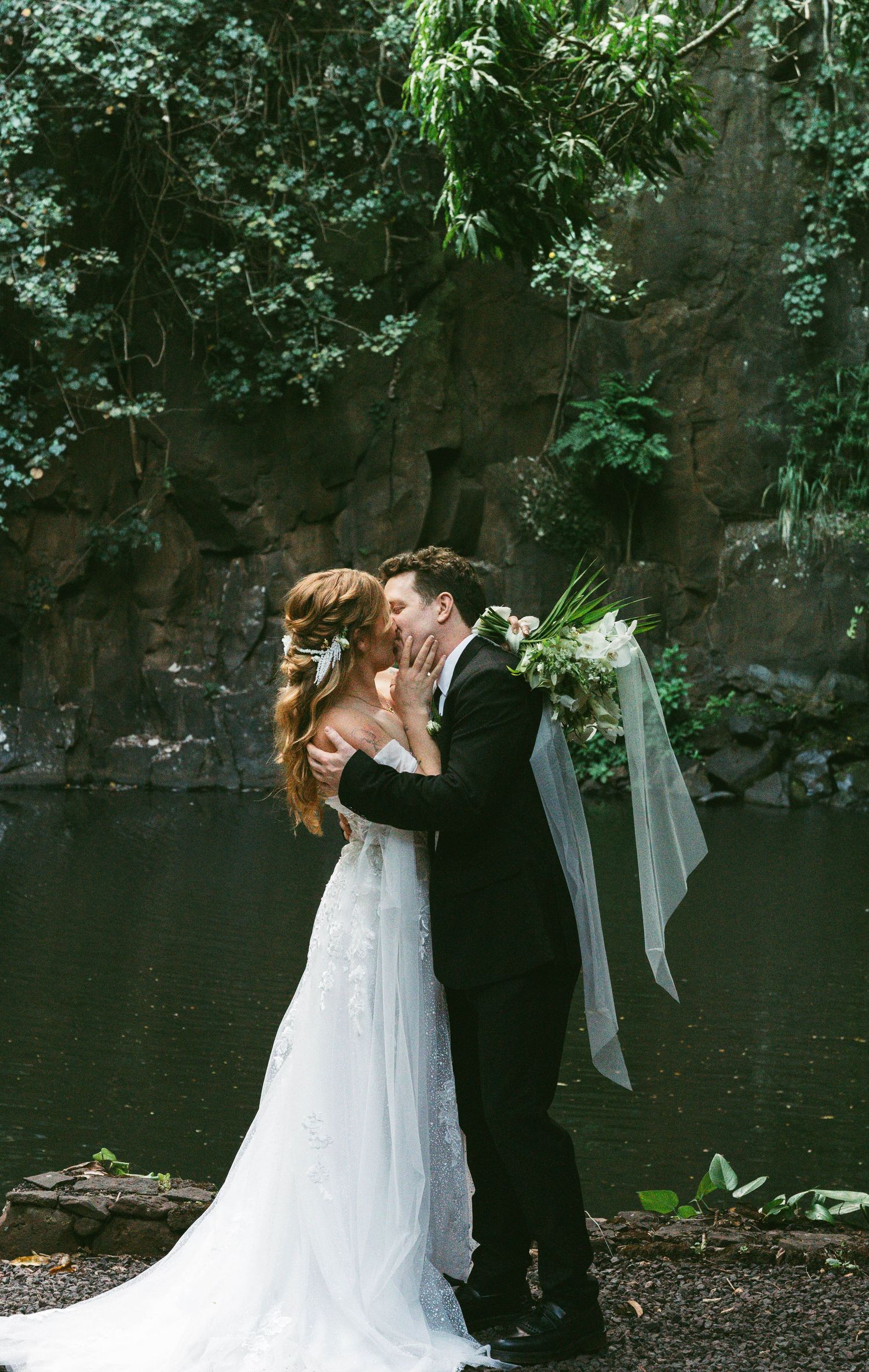 Couple in wedding attire embracing by tranquil pond with waterfall and greenery.