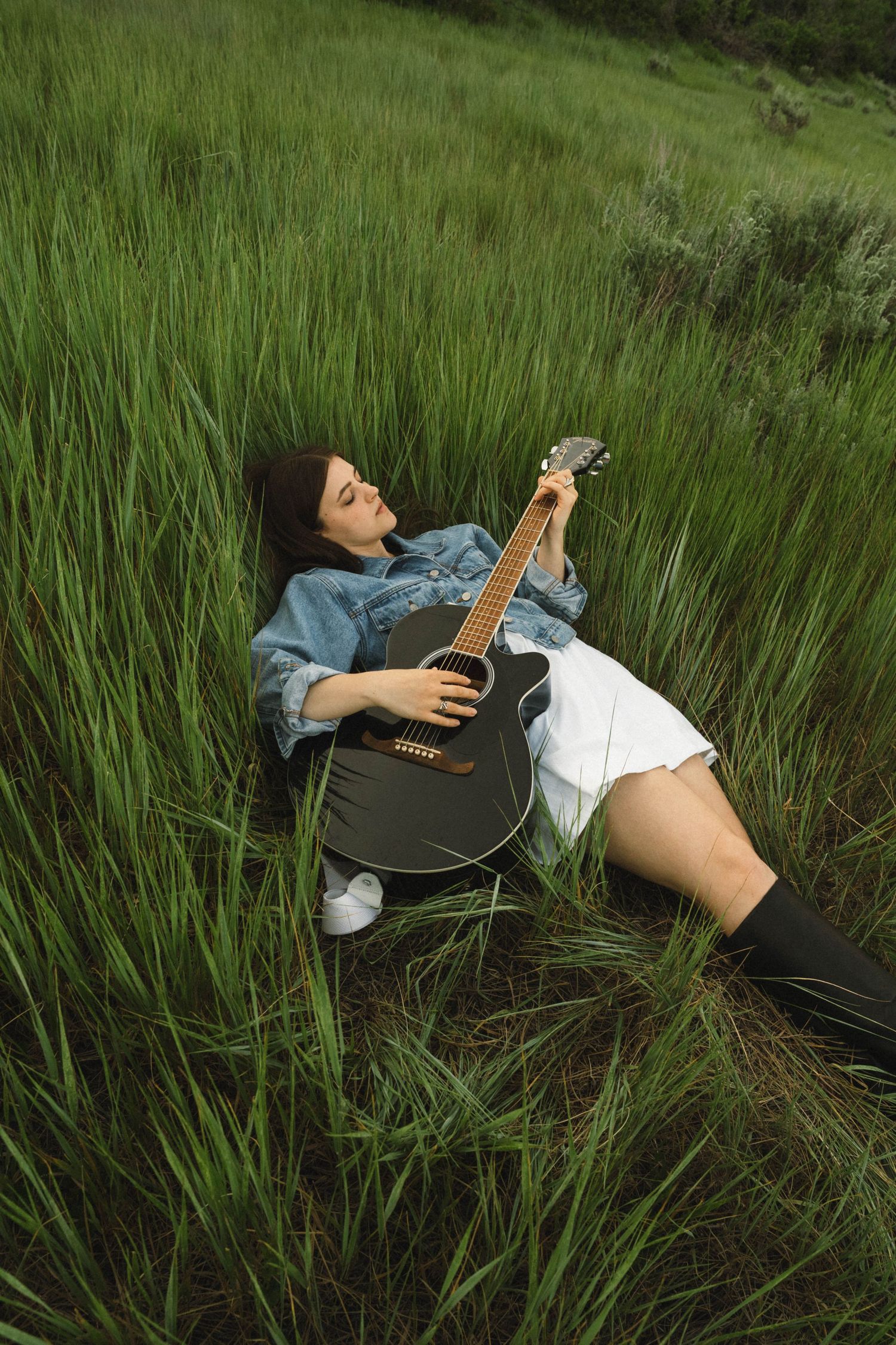 Young woman lying in tall green grass playing a black acoustic guitar, wearing a denim jacket and white skirt.