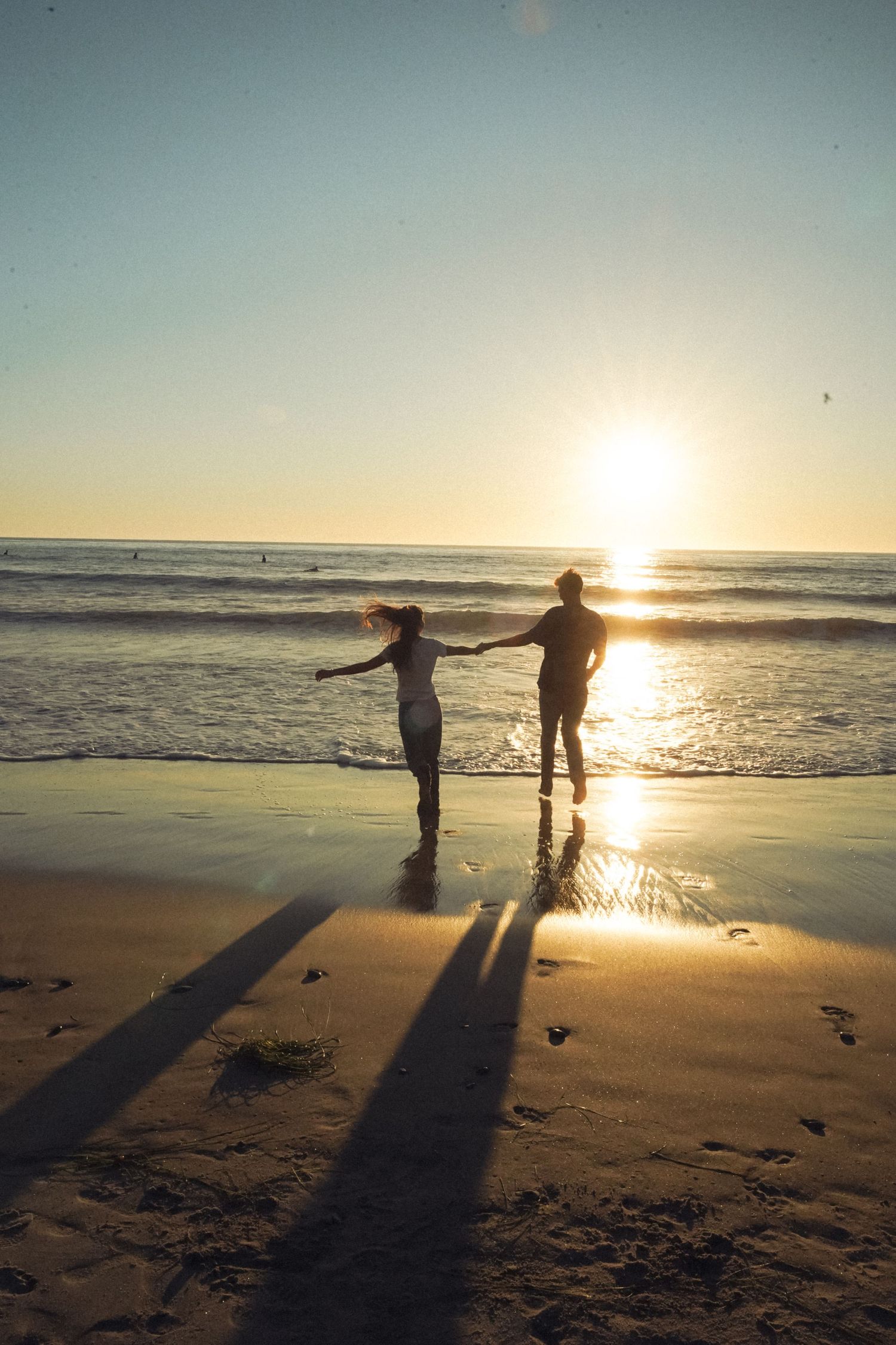 Silhouettes of two people casting long shadows on a Californian beach at sunset while standing near the ocean waves.