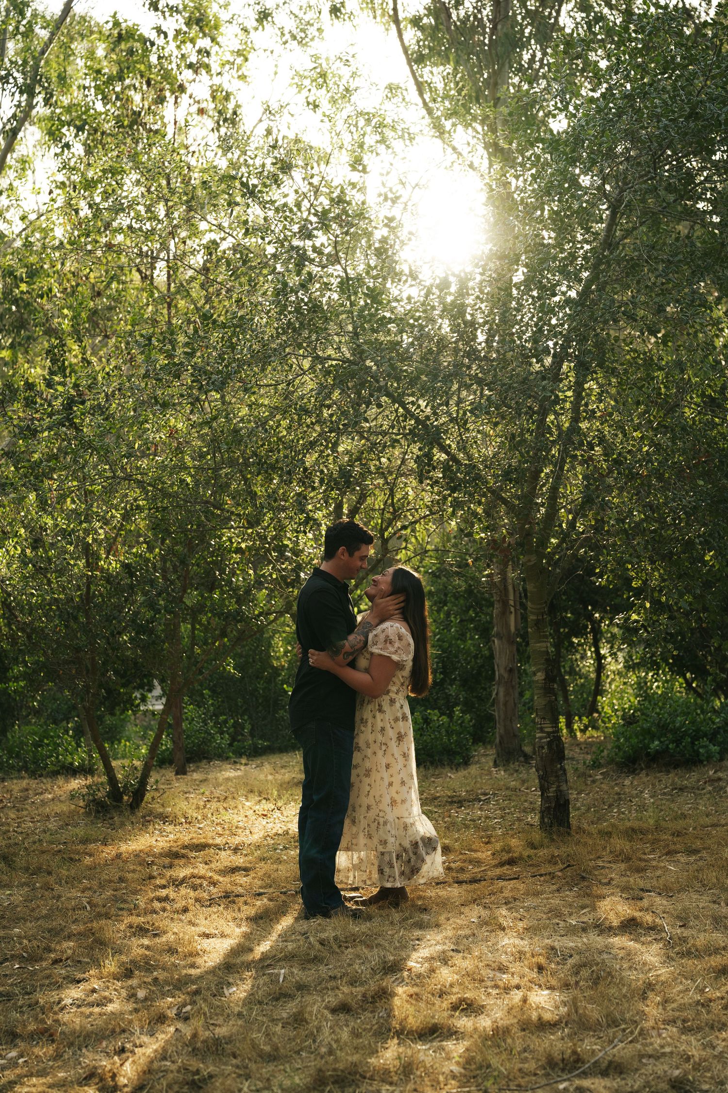 Couple embracing face to face in sun-drenched forest clearing with warm golden backlight through trees.