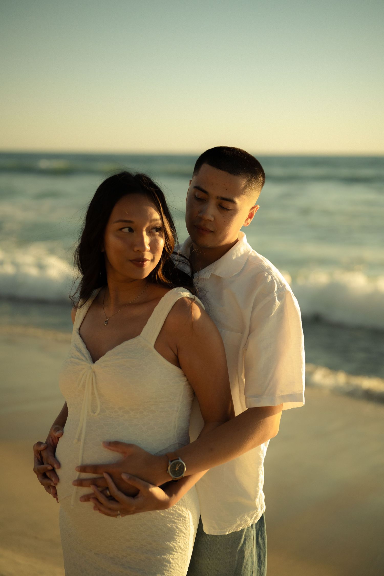Expecting couple embracing on a beach at sunset, man in white shirt holding pregnant woman's belly by the ocean.