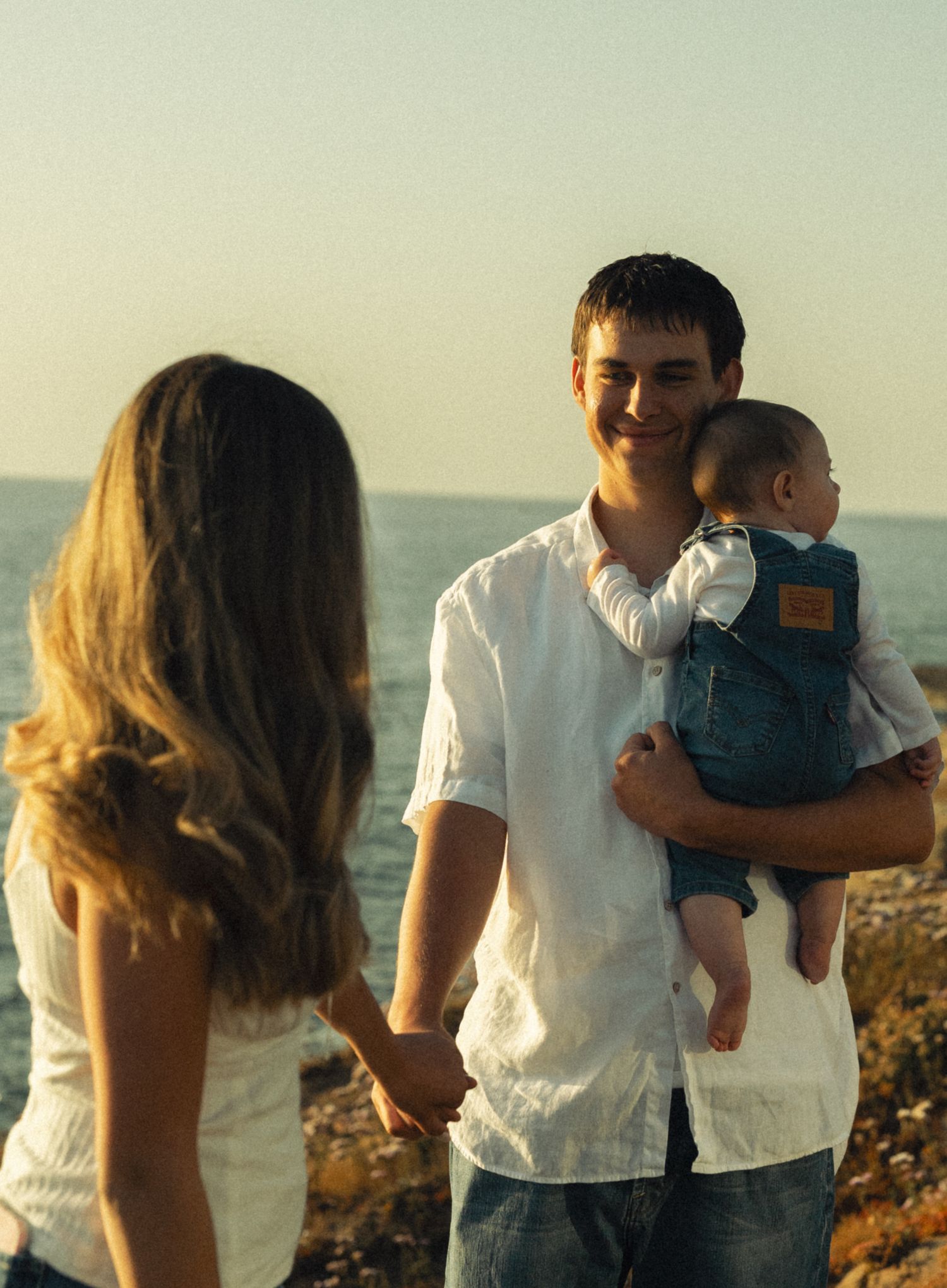 Young couple holding hands near ocean shore, man in white shirt smiling while holding baby in denim carrier at sunset.