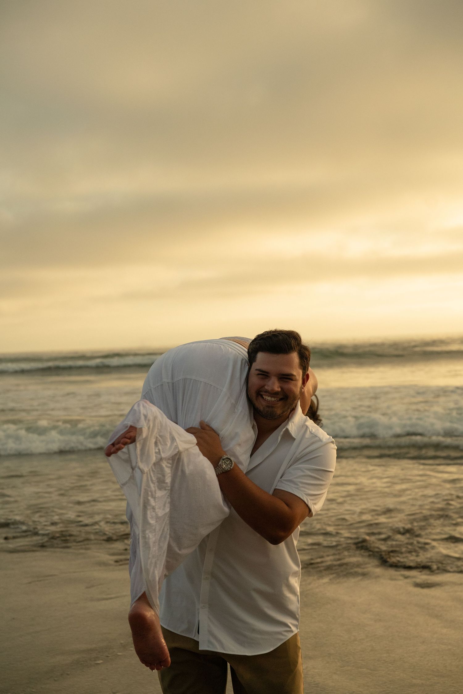 Smiling man carrying woman over his shoulder on a sandy beach during a warm golden sunset.