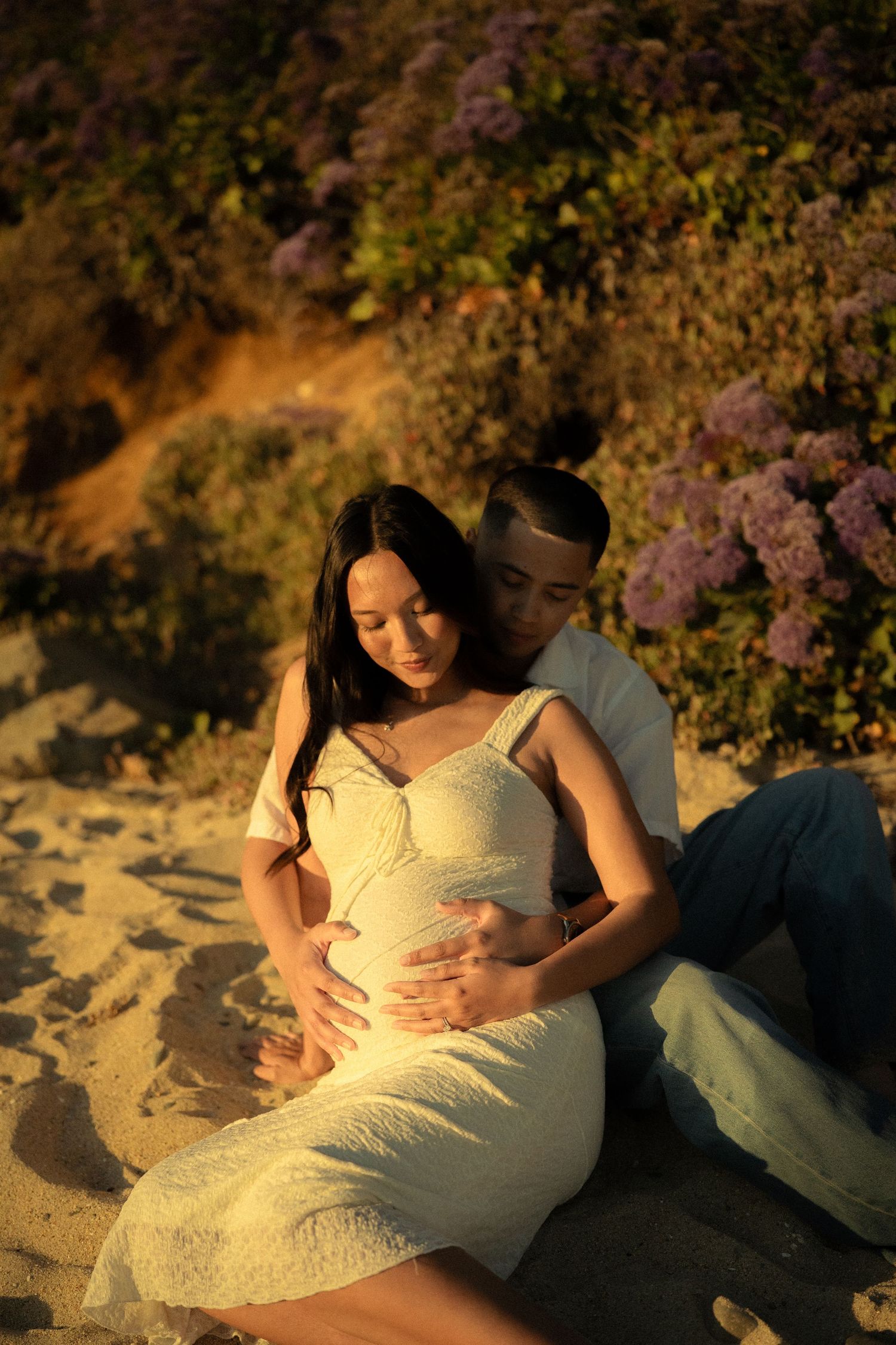 Pregnant couple sitting on sandy beach at golden hour, man embracing woman in white lace dress with purple flowers behind.
