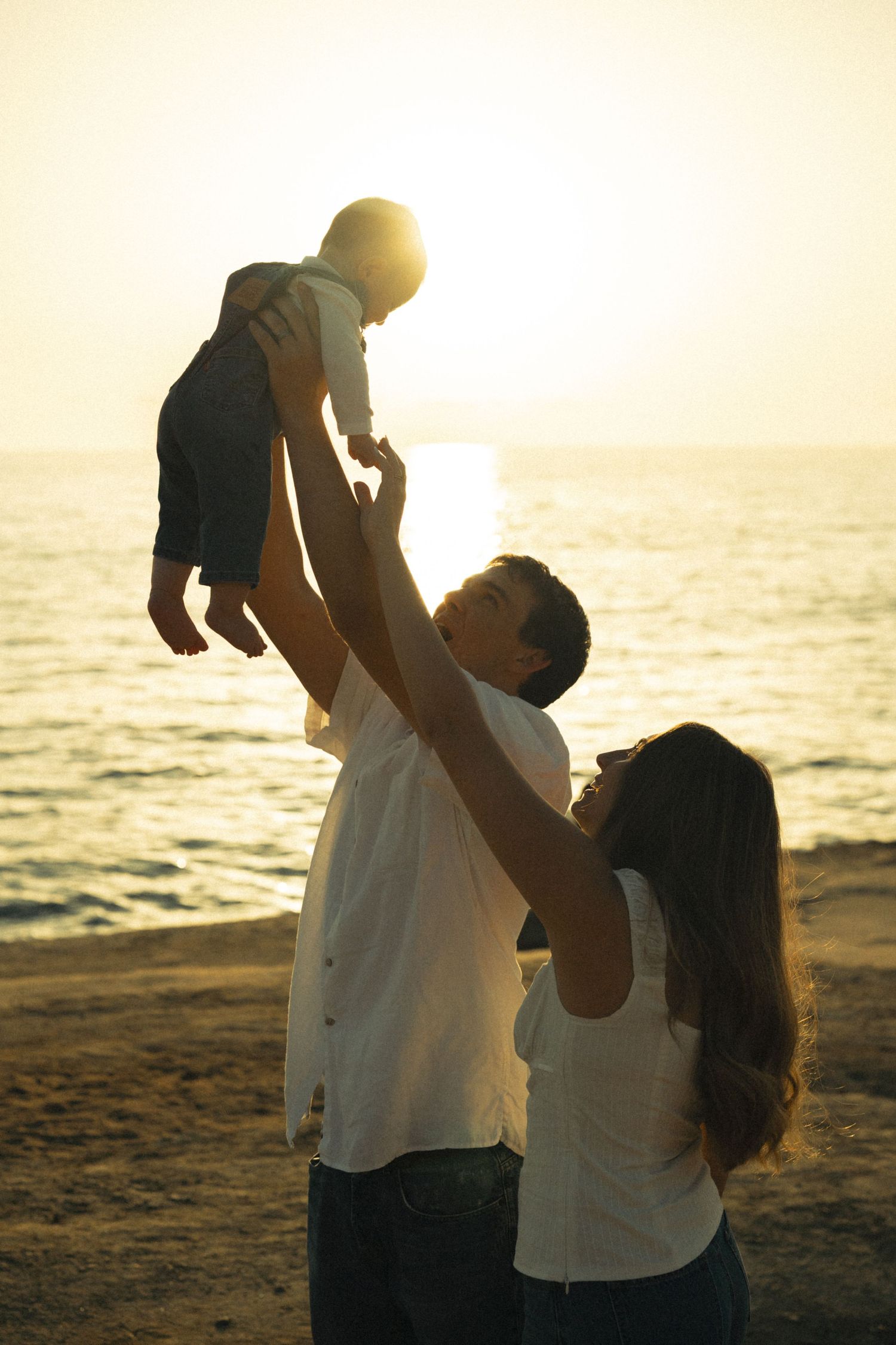 Parents lifting baby at sunset beach, golden backlit silhouette of happy family enjoying ocean shore moment.