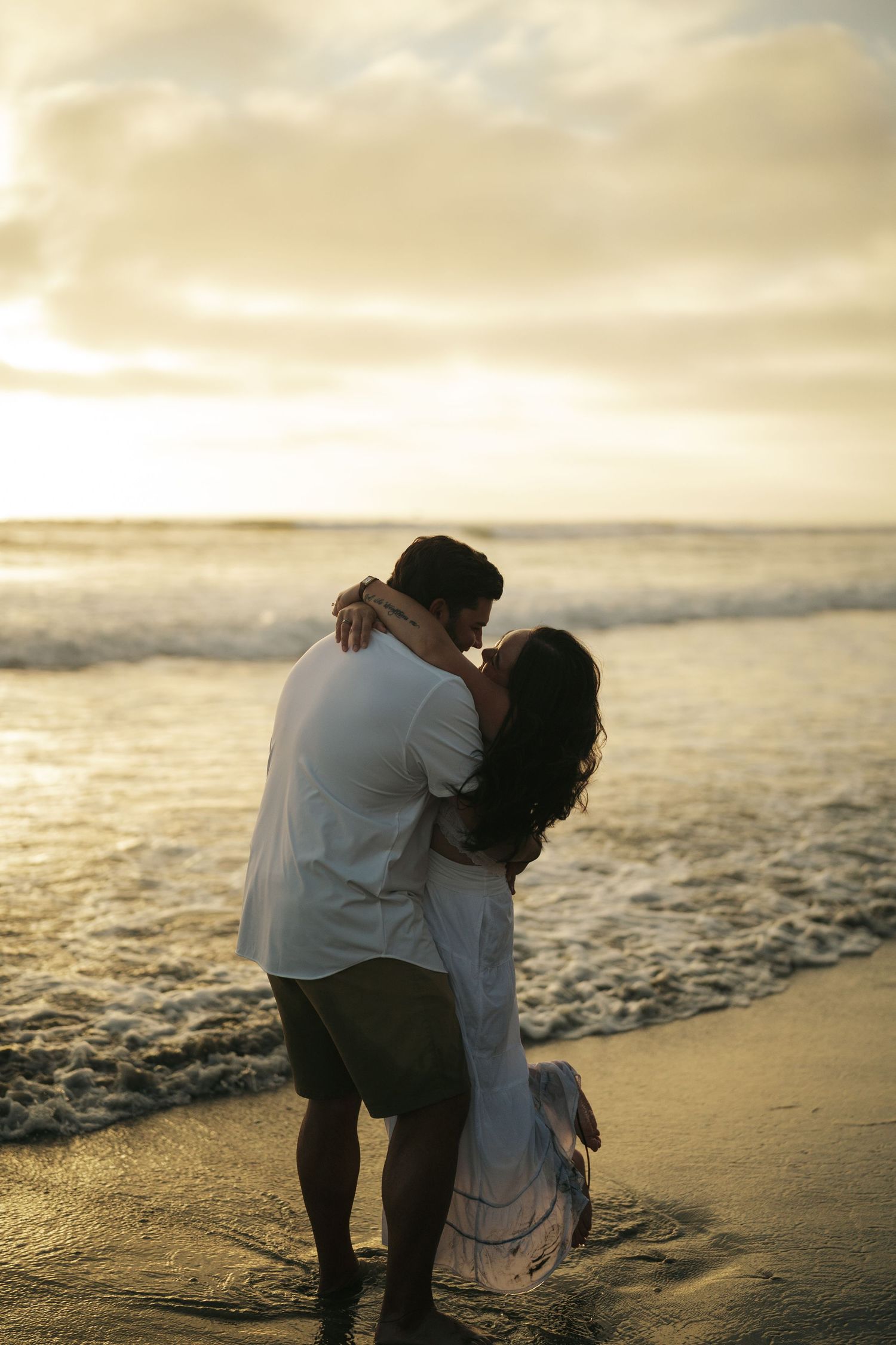 Couple sharing a romantic kiss on the beach at golden sunset, waves washing over their feet.