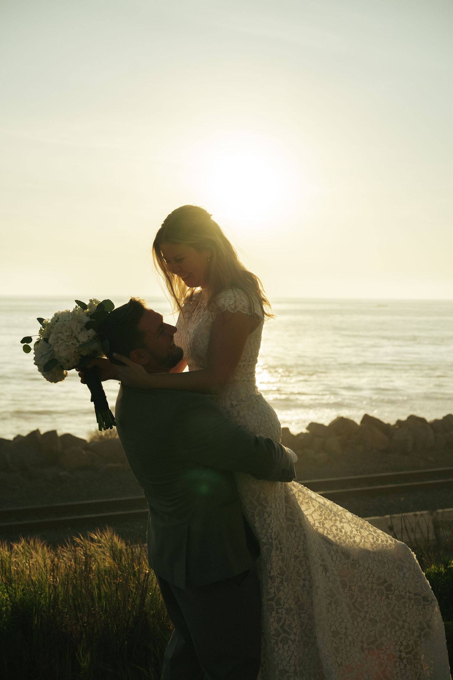 Groom lifts bride in silhouette against glowing sunset over the ocean during a romantic beach wedding.