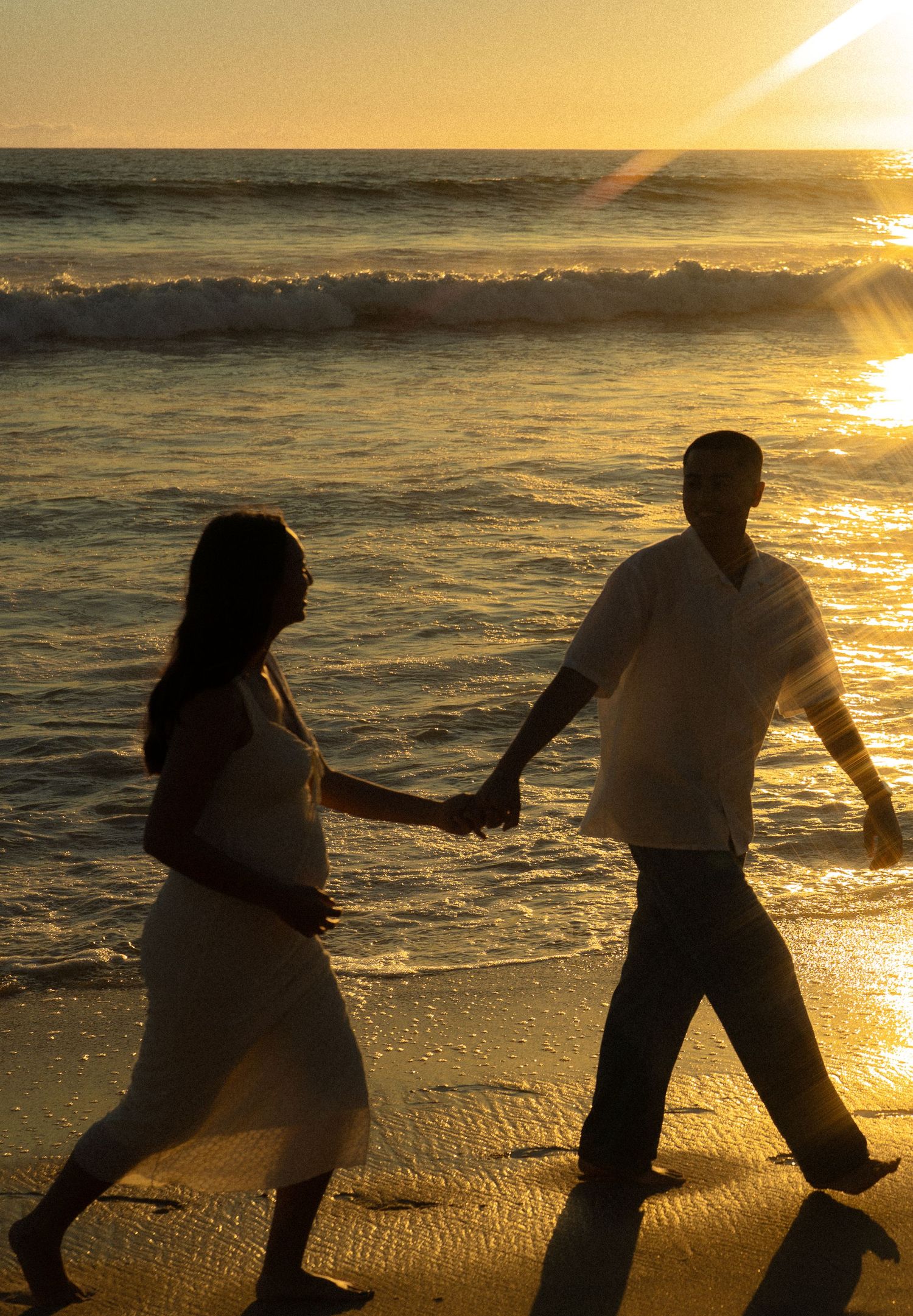 Pregnant couple silhouetted walking hand in hand along beach at golden sunset.