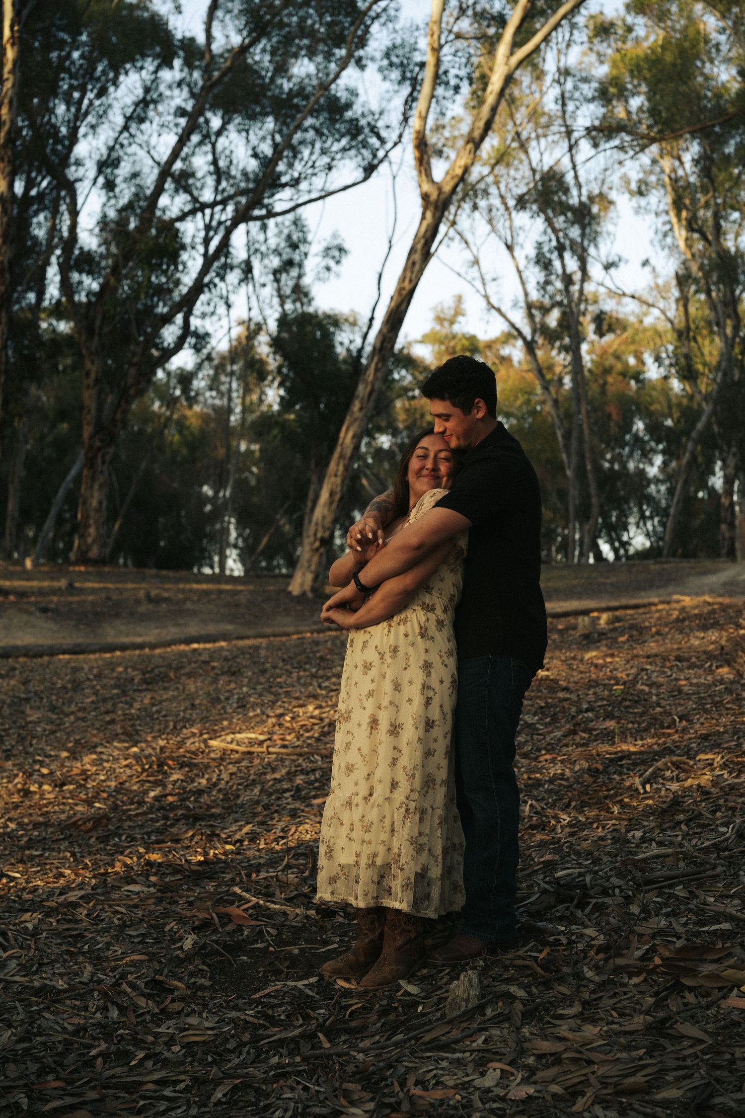 Couple embracing warmly in a golden-lit eucalyptus forest, woman in floral dress smiling contentedly.