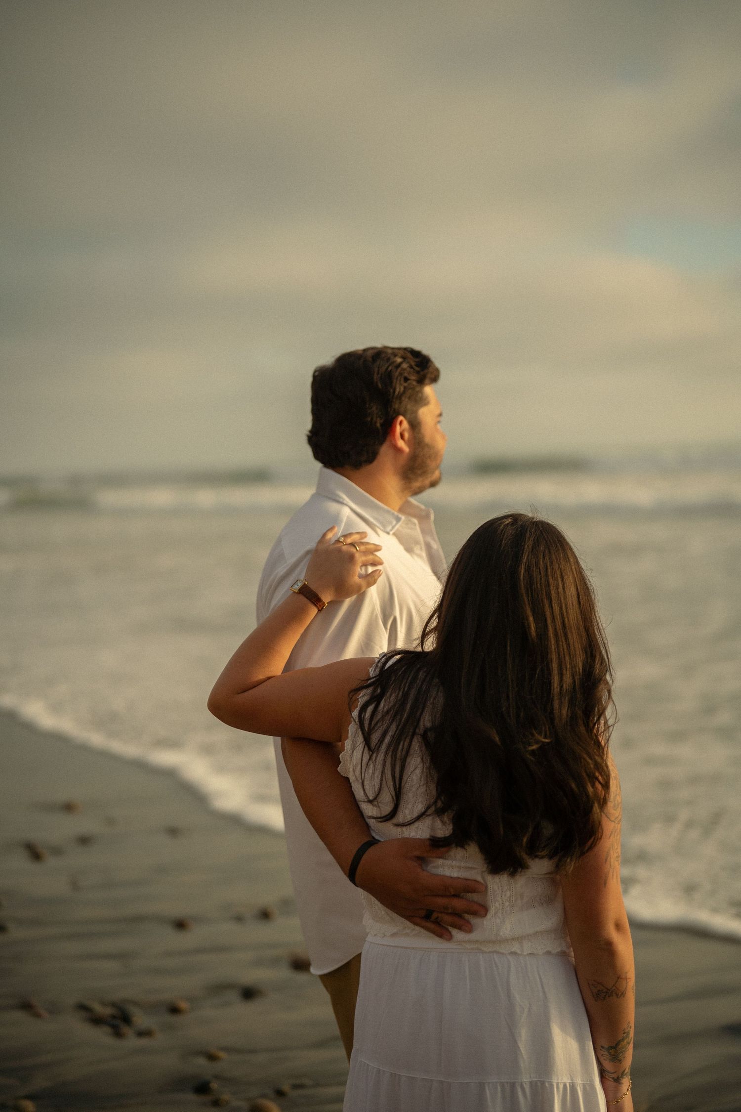 Couple embracing on a sandy beach at golden hour, gazing at ocean waves in matching white outfits.