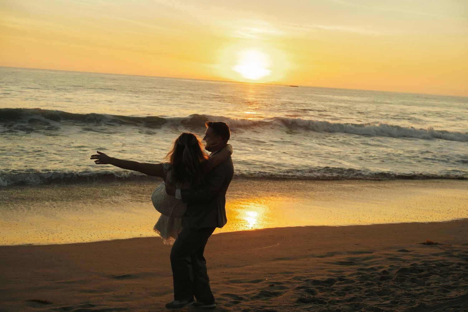 Newlyweds dance joyfully on sandy beach as brilliant golden sunset reflects over ocean waves behind them.