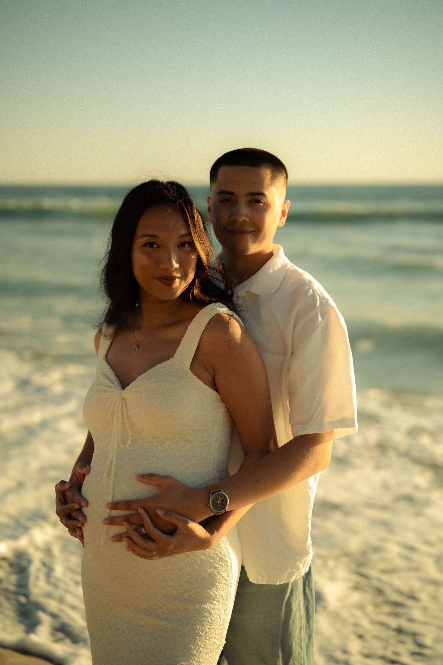 Expecting couple embracing on beach at sunset, man holding pregnant woman's belly.