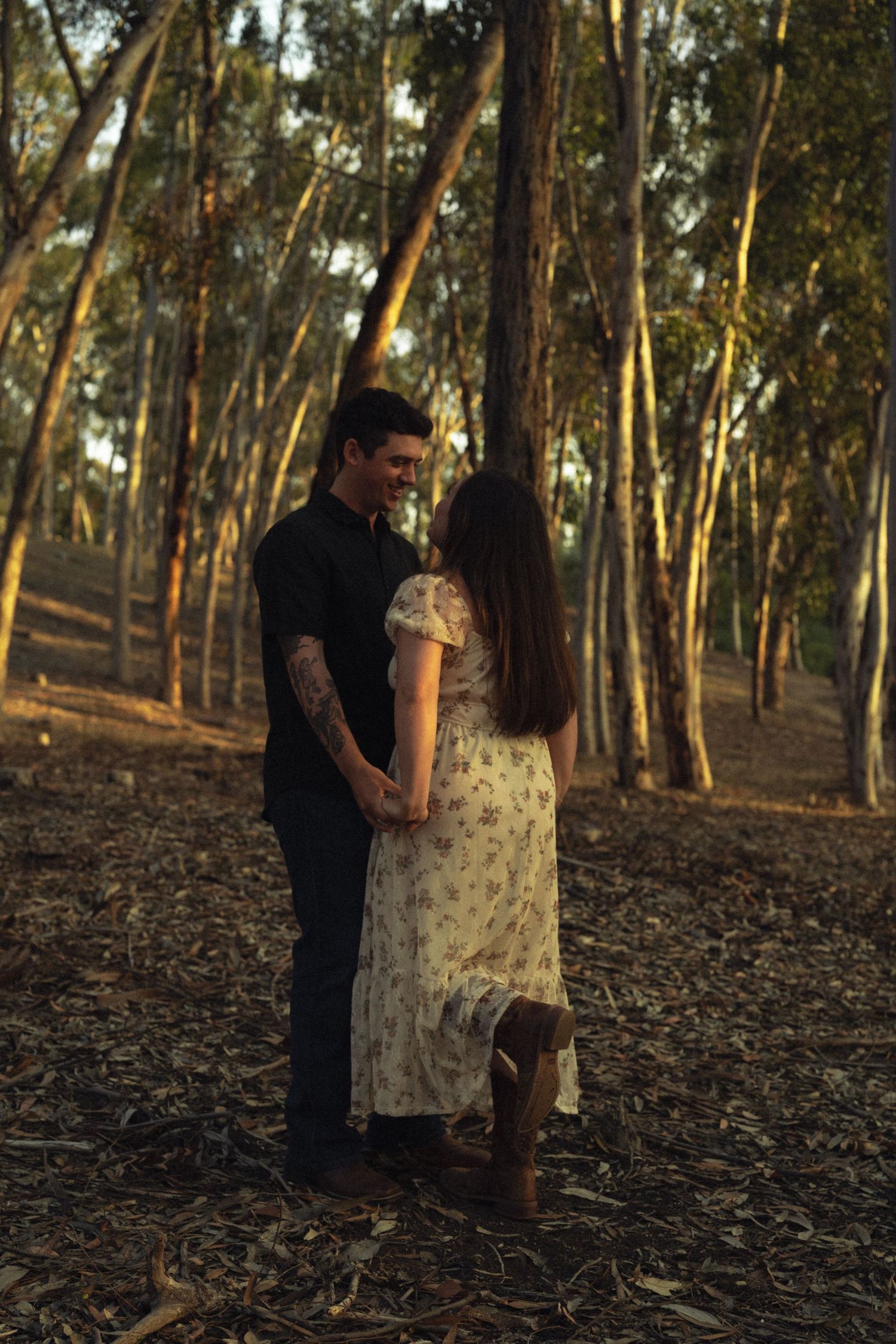 Couple holding hands and smiling at each other in a golden-hour eucalyptus forest setting.