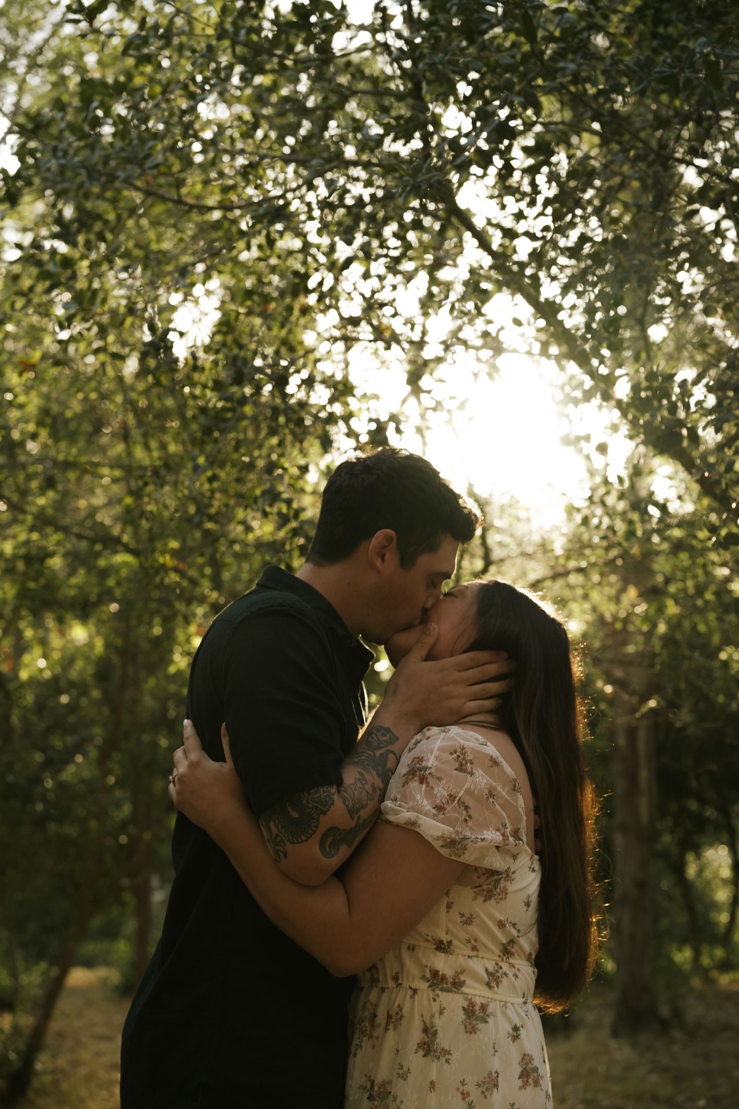 Couple sharing a romantic kiss outdoors under lush green trees with golden sunlight filtering through the leaves.