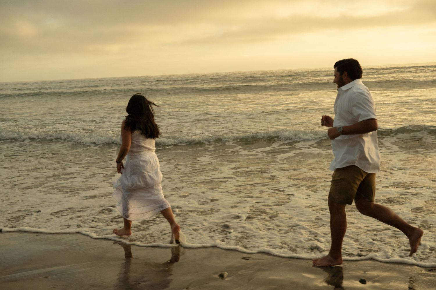 A couple runs playfully along a sandy beach shore together during a golden sunset.