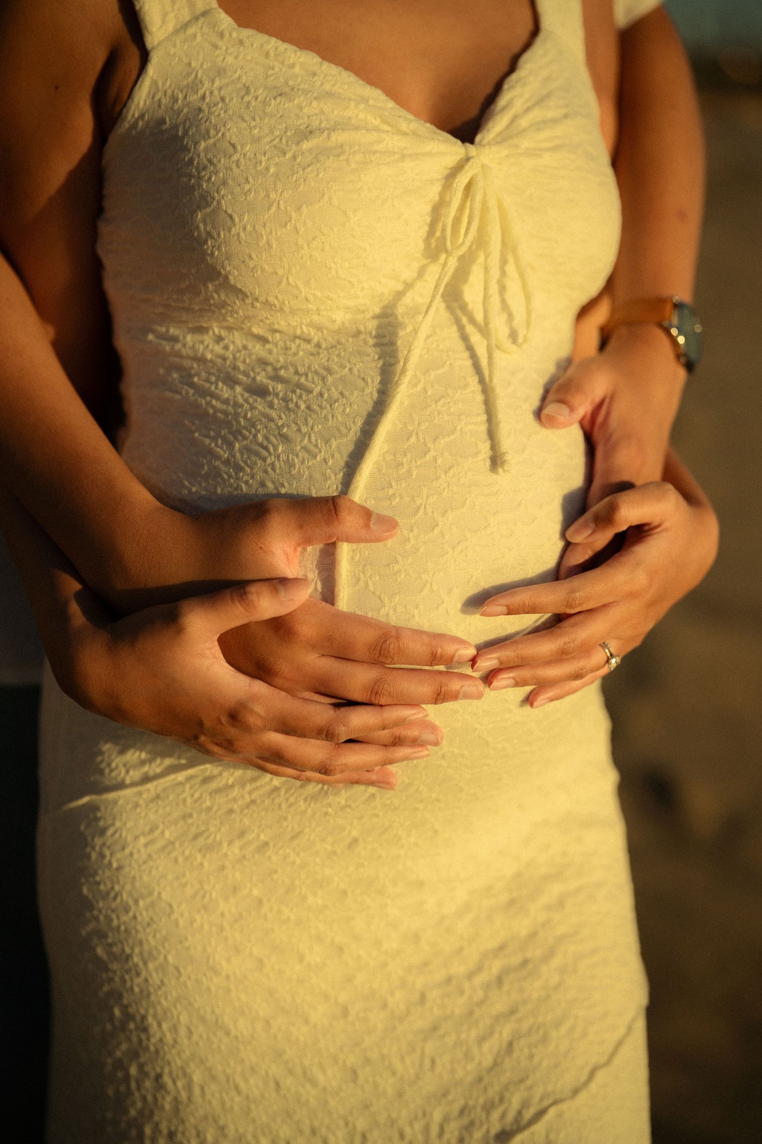 Close-up of couple's hands cradling pregnant belly in white lace dress at sunset.