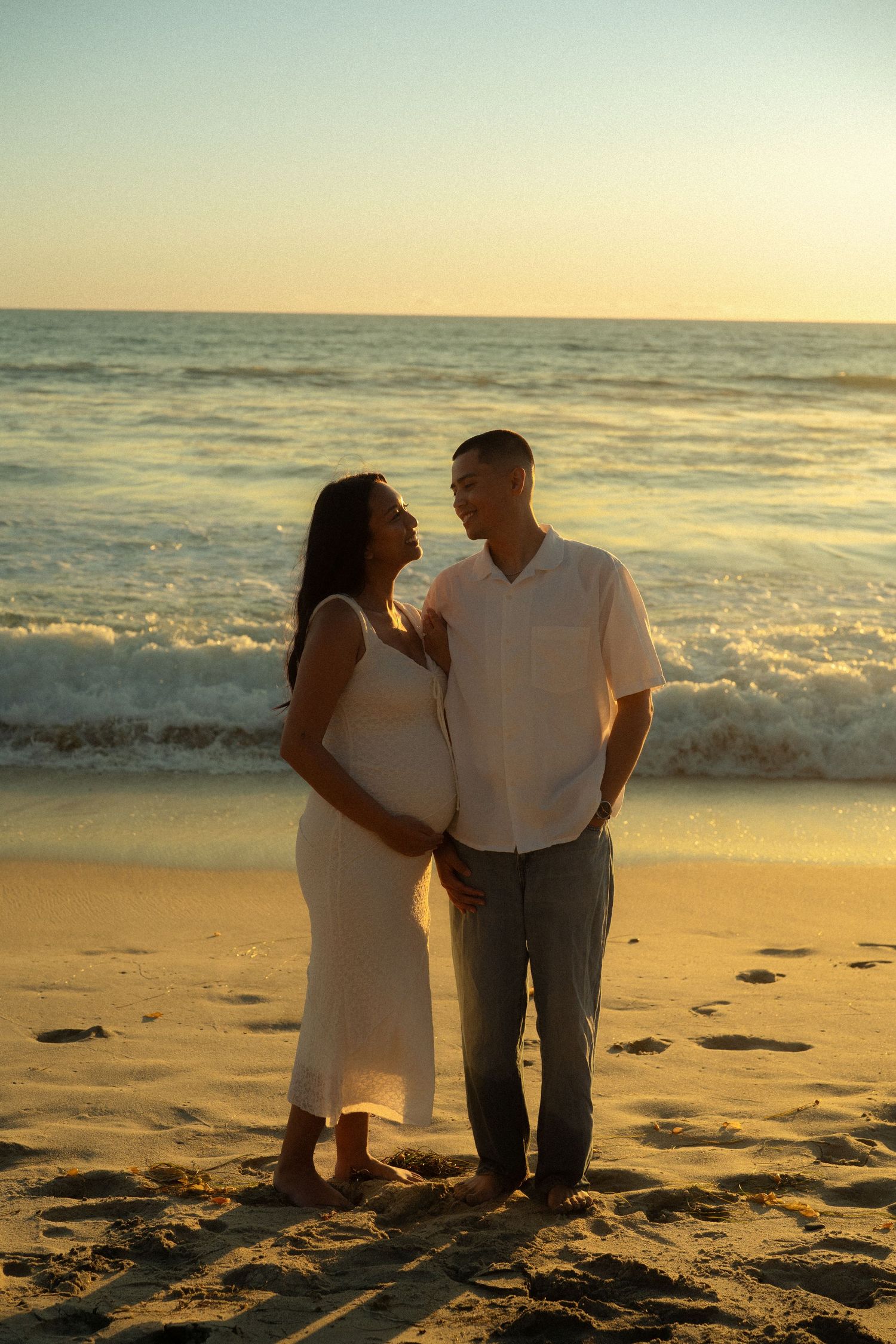 Happy expecting couple standing face to face on sandy beach during warm golden sunset.