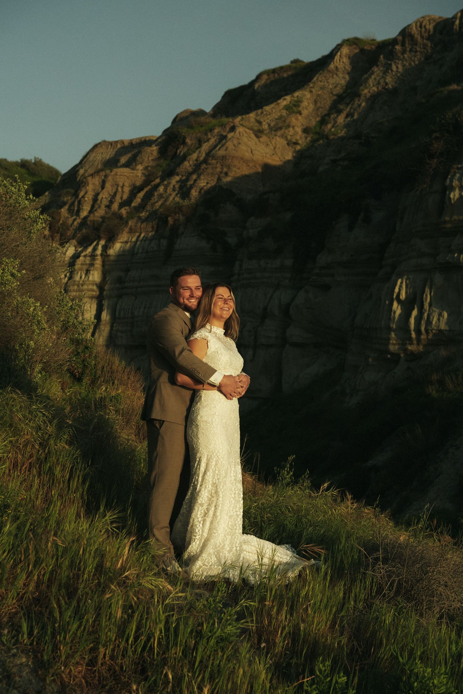 Couple embracing in wedding attire on grassy hillside with rocky cliffs.