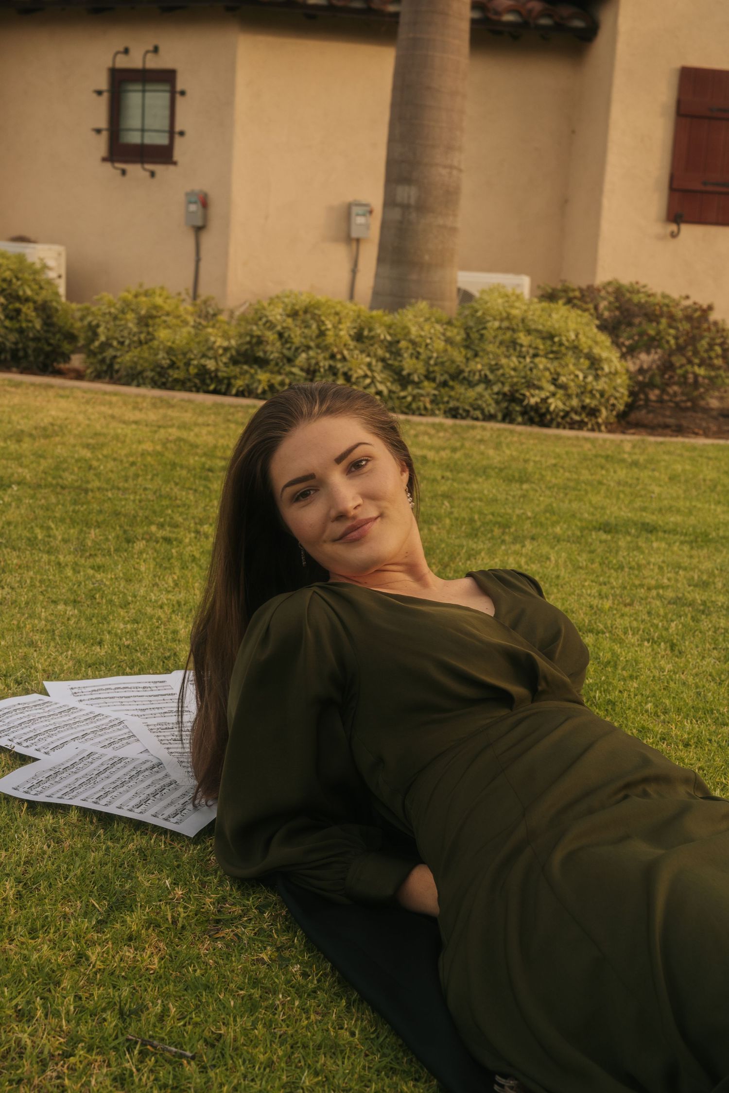 Young woman in olive green dress lying on grass with sheet music, relaxing outdoors near a building.