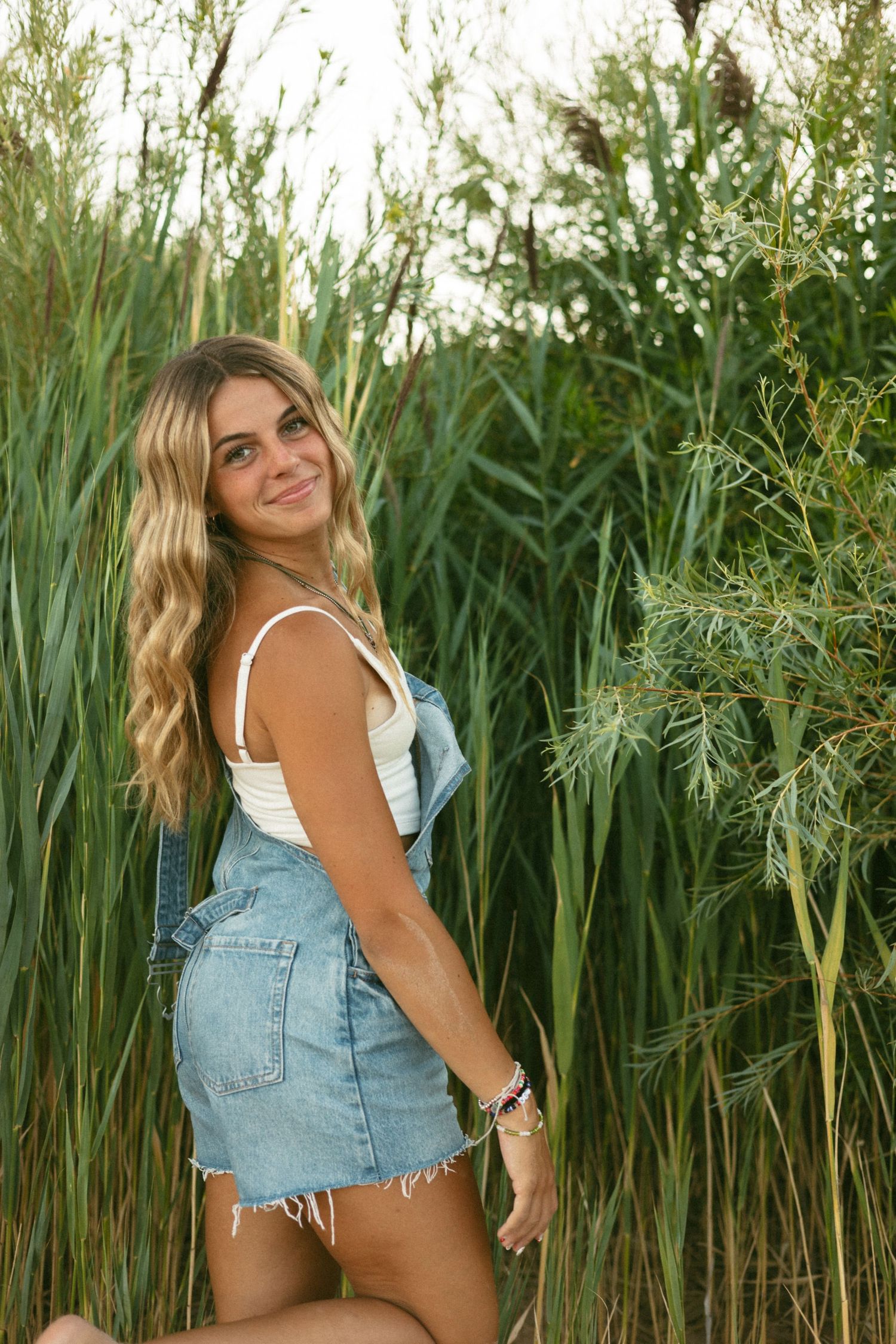Young blonde woman in denim overalls smiling among tall green reeds outdoors.