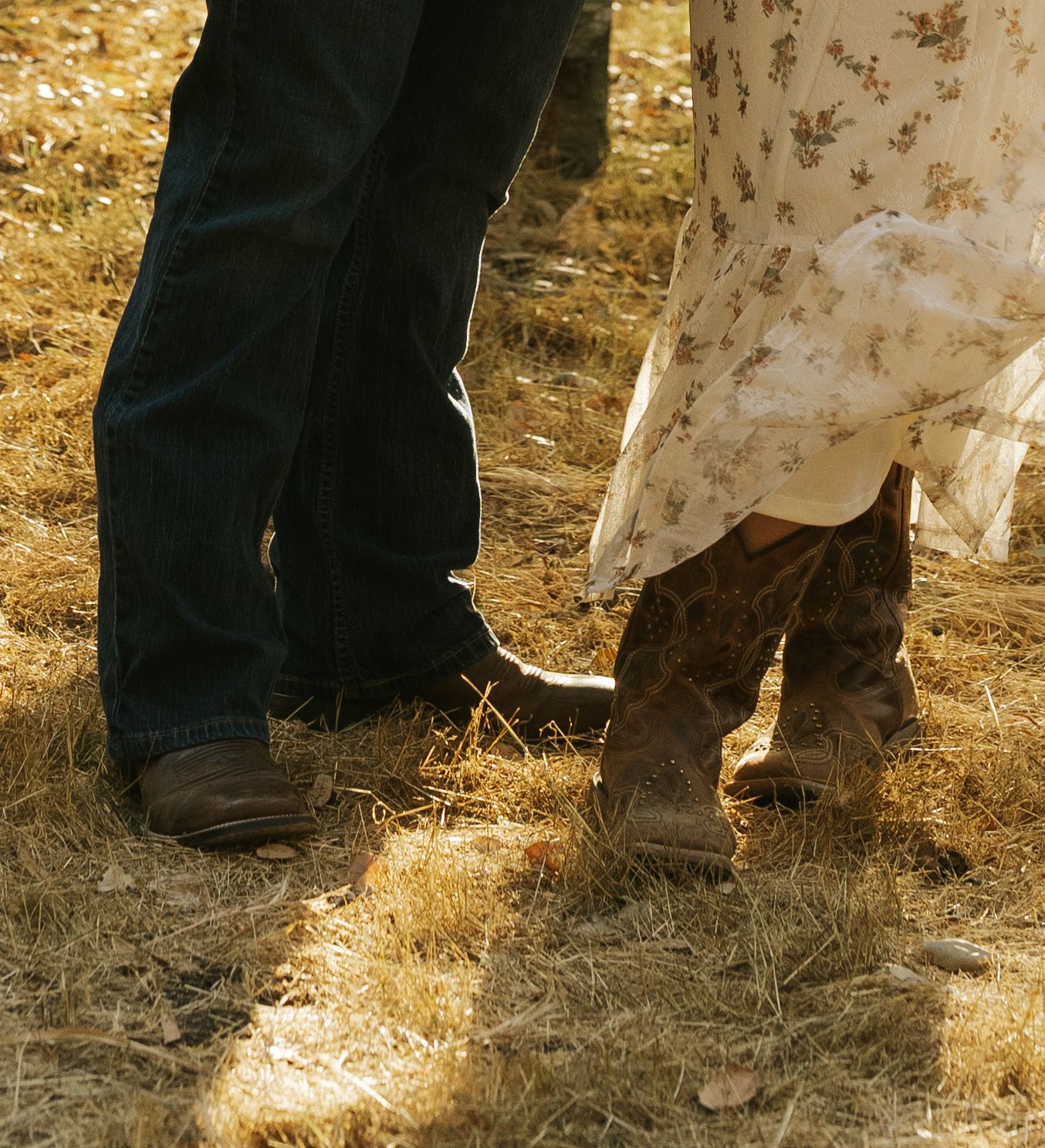 Couple wearing cowboy boots and jeans with floral dress standing on dry grass field.
