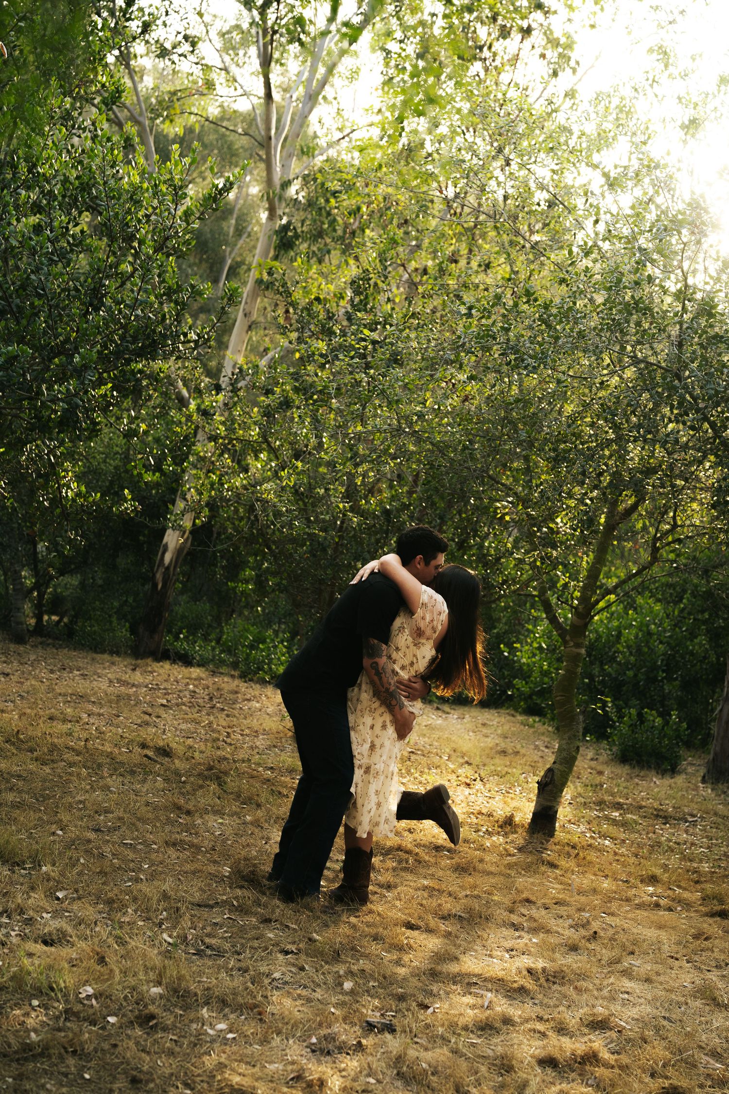 Couple sharing a romantic dip kiss in a bright sunlit green tree-filled outdoor setting.