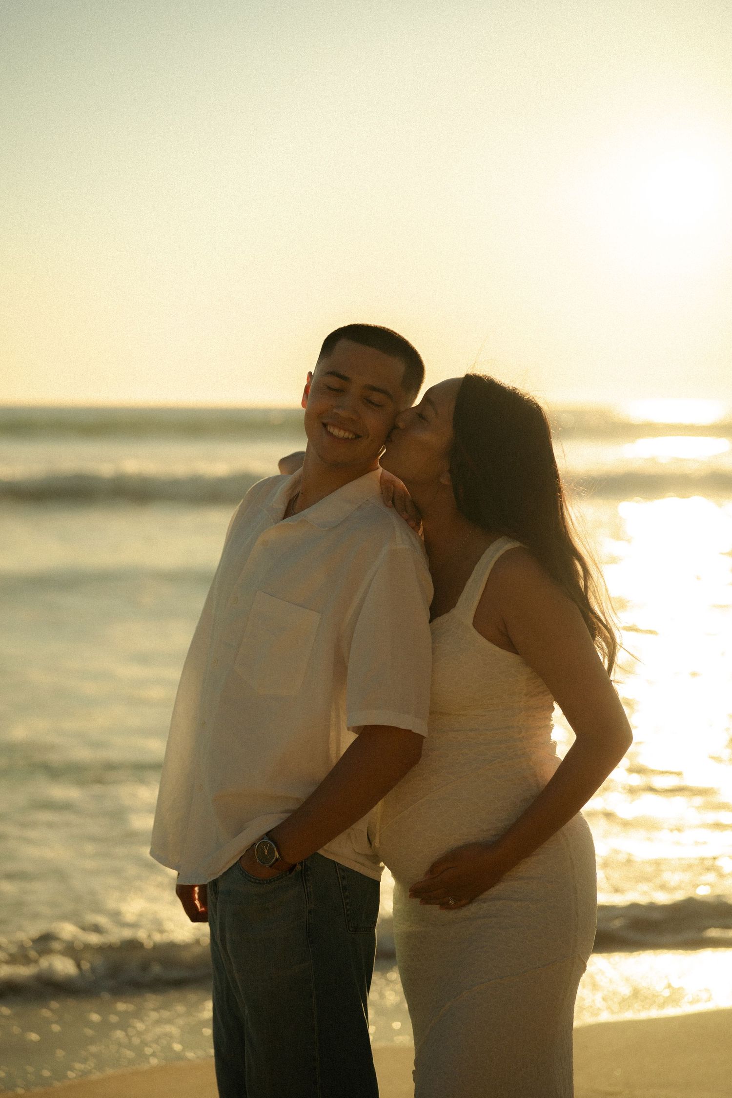 Pregnant woman in white dress kissing smiling man on cheek during golden hour beach sunset.