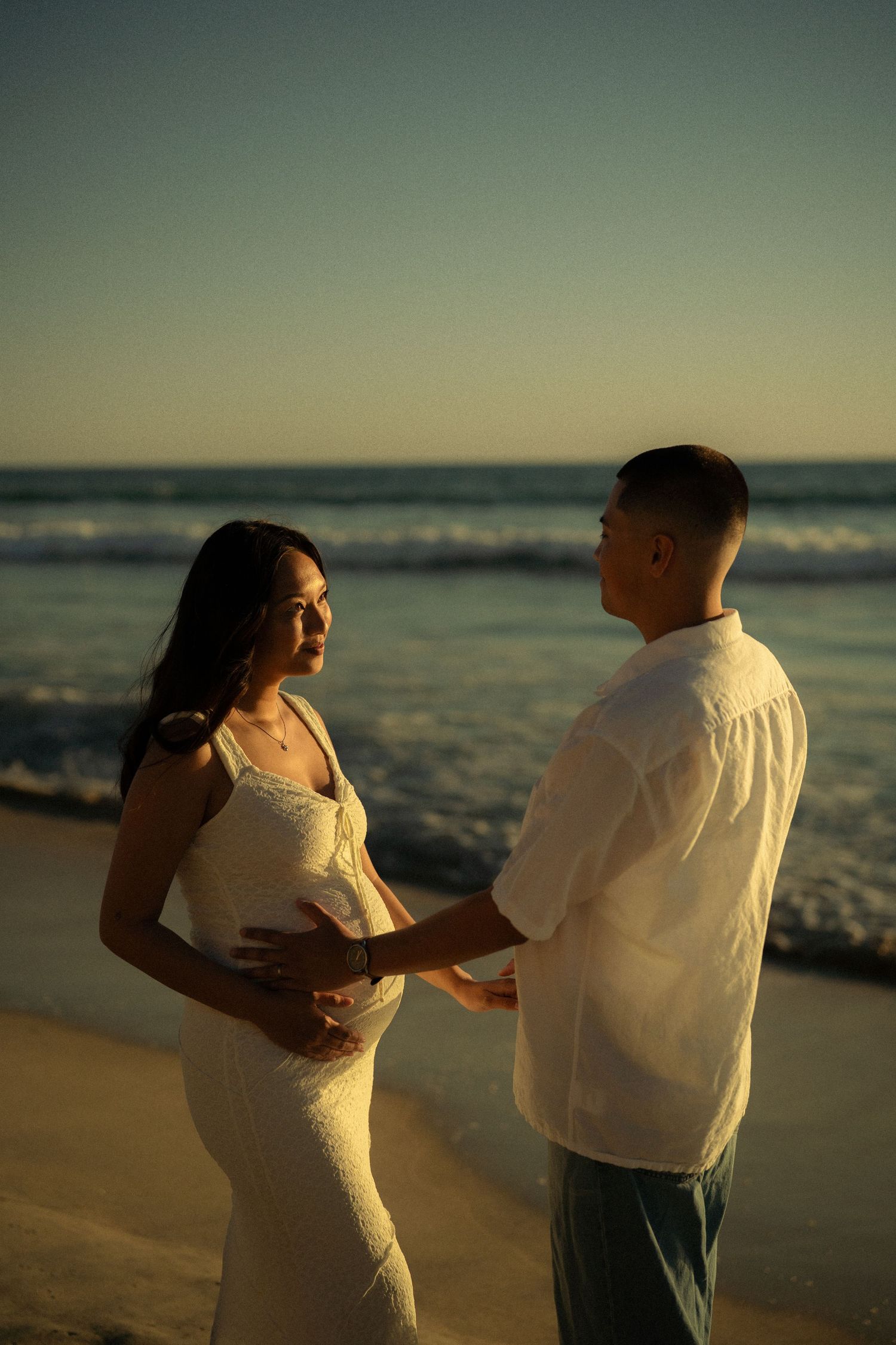 Couple sharing tender moment on beach at sunset, pregnant woman in white dress held by partner near ocean waves.