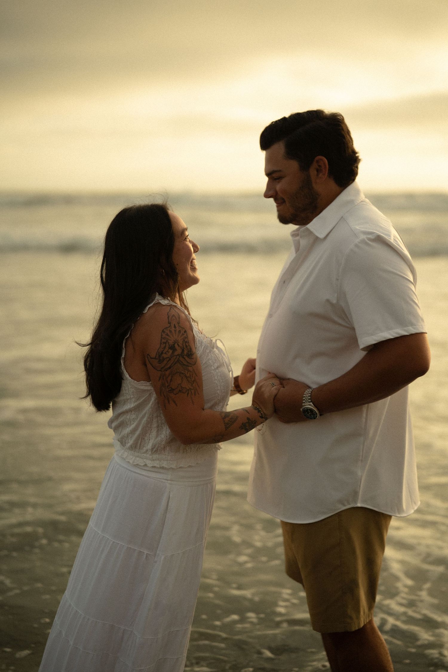 Couple in white outfits holding hands on beach at golden sunset, woman with arm tattoo smiling at man.