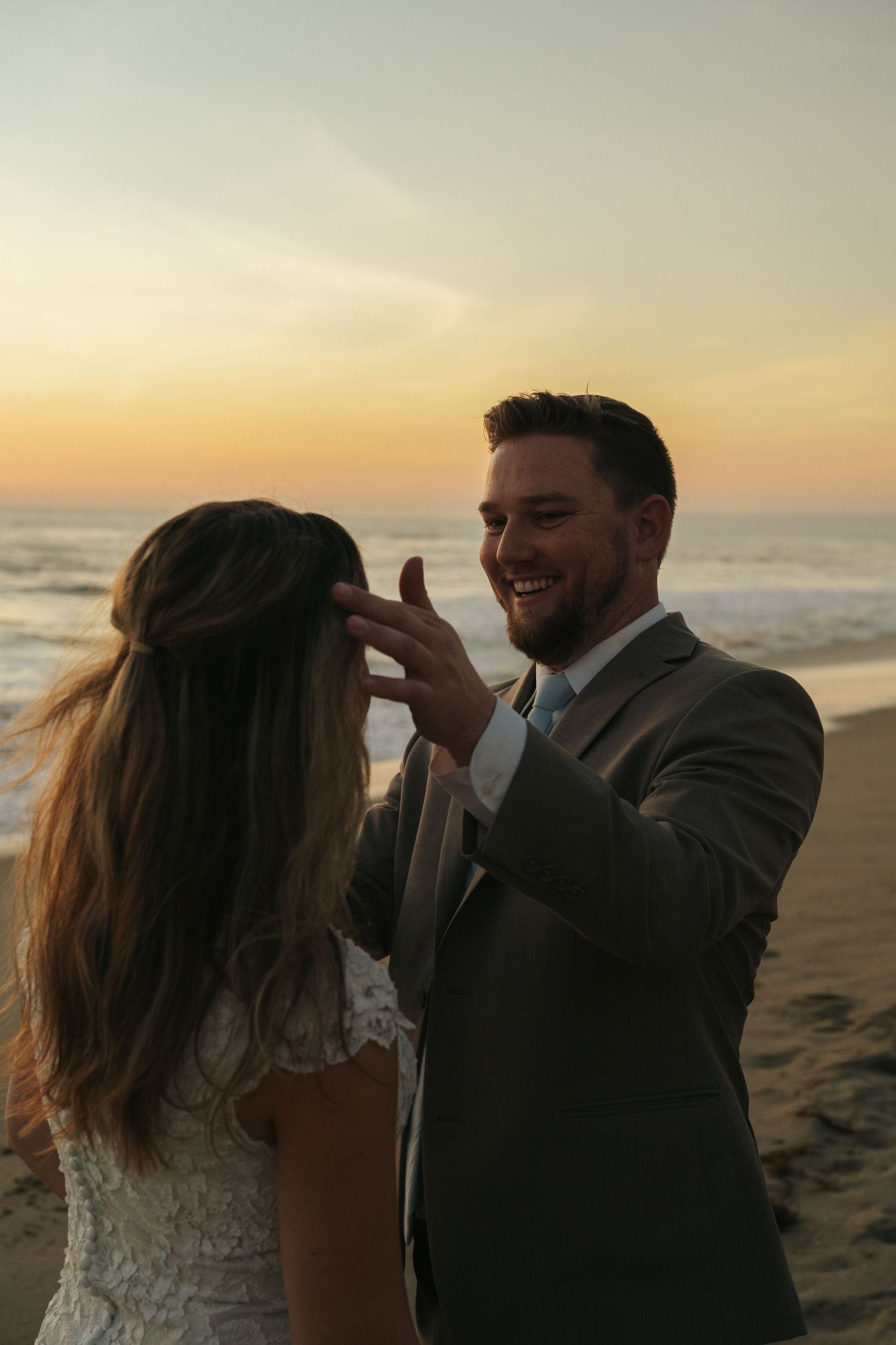 Smiling groom tenderly brushing brides hair on sandy beach at sunset by the ocean.