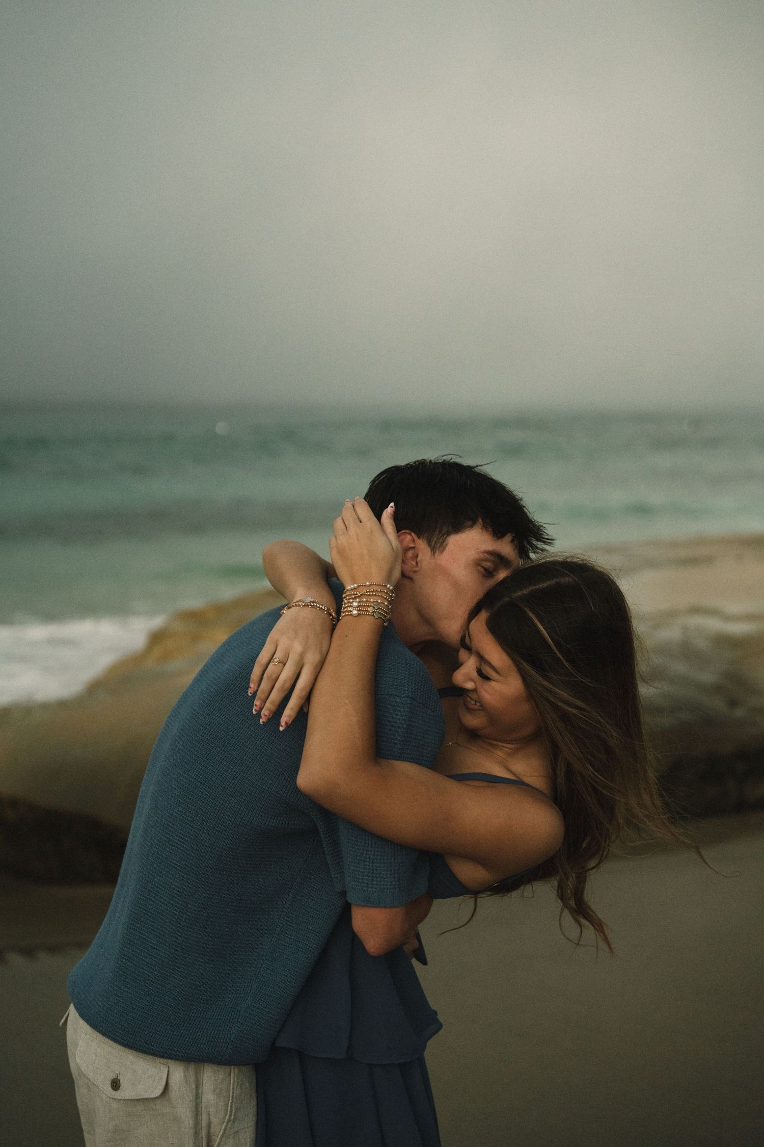 Couple sharing a romantic embrace and kiss on a moody beach with ocean waves in the background.