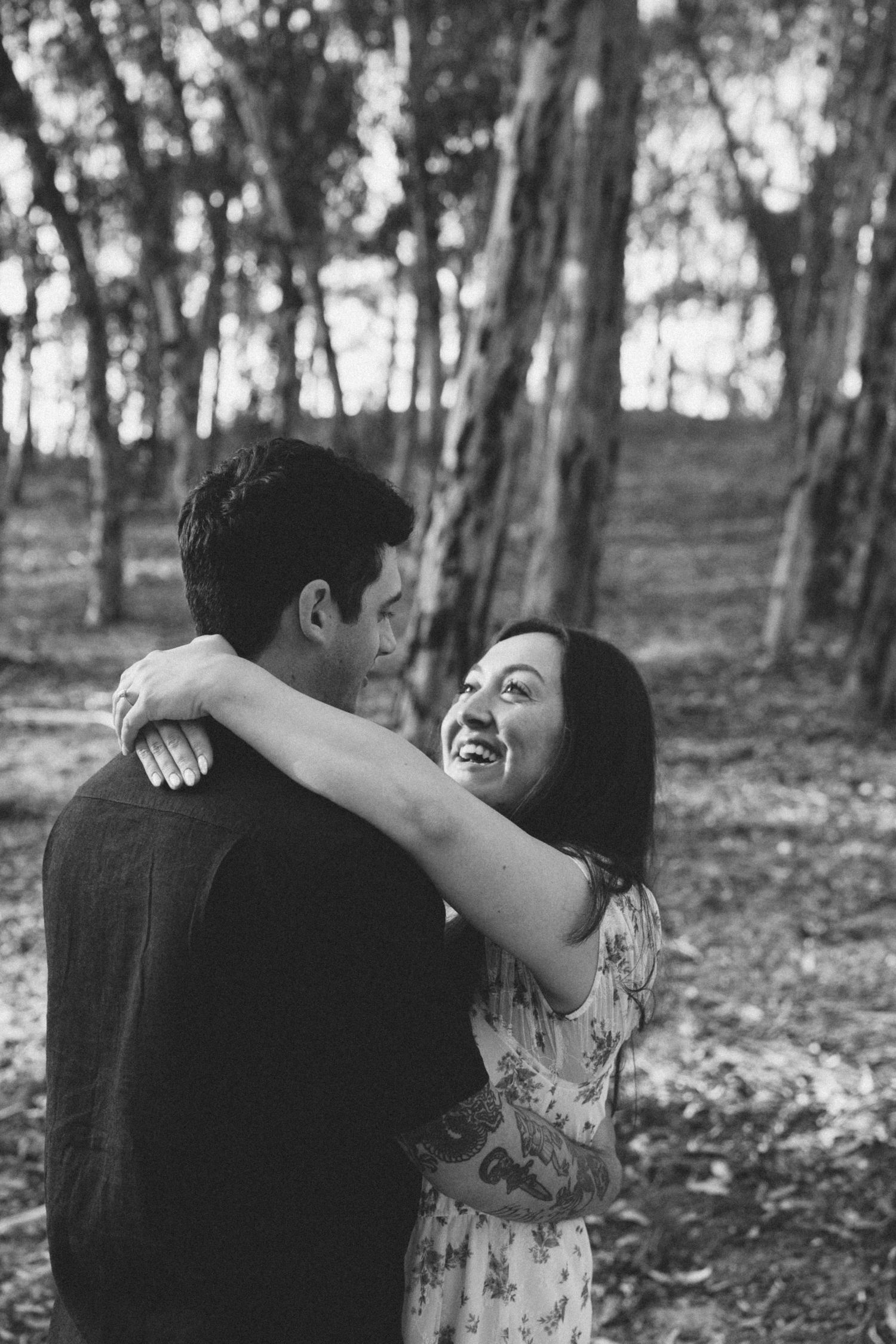 Black and white photo of a laughing woman embracing a man among tall trees in a serene forest setting.