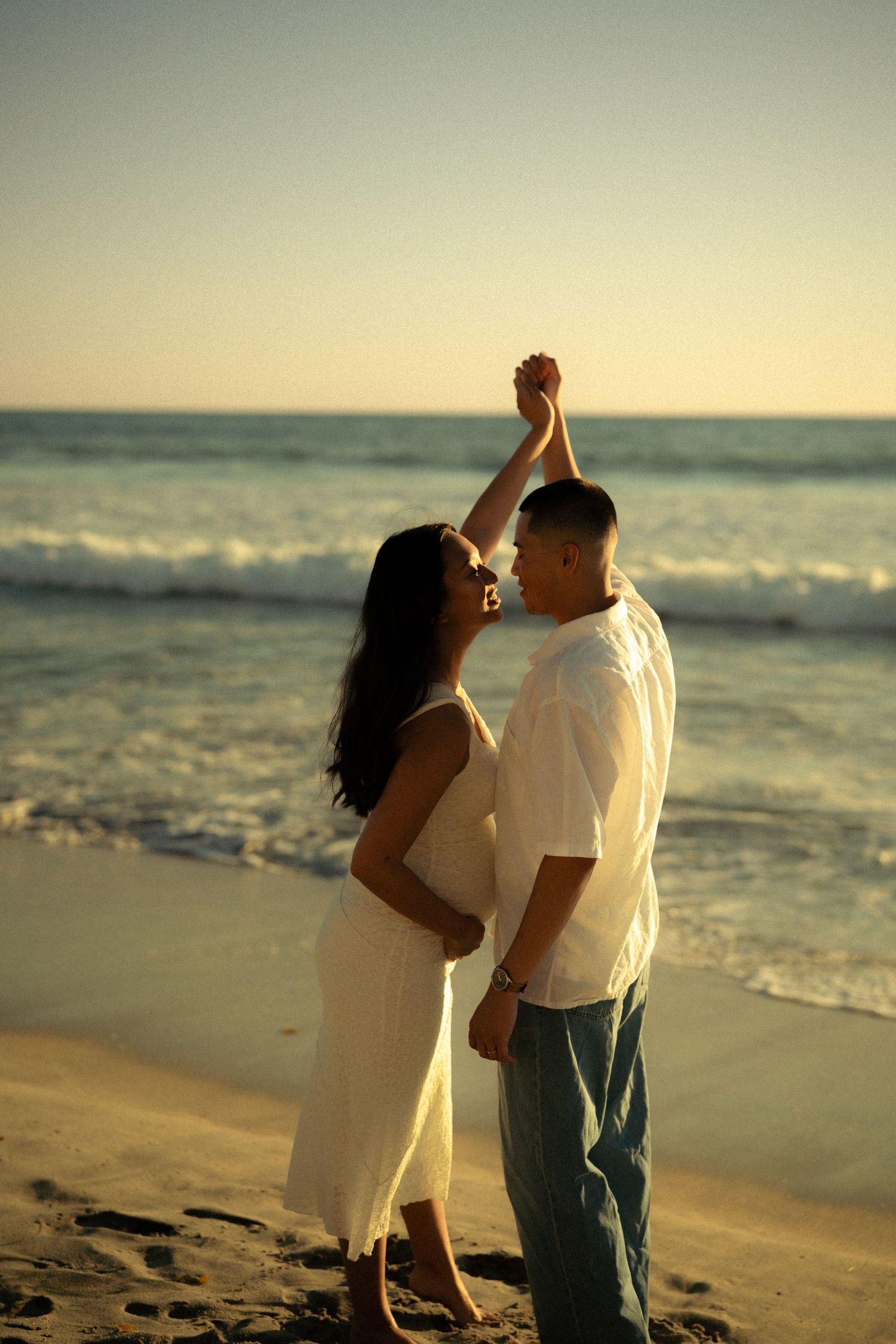 Romantic couple dancing on a sandy beach at sunset, holding hands with ocean waves in the background.