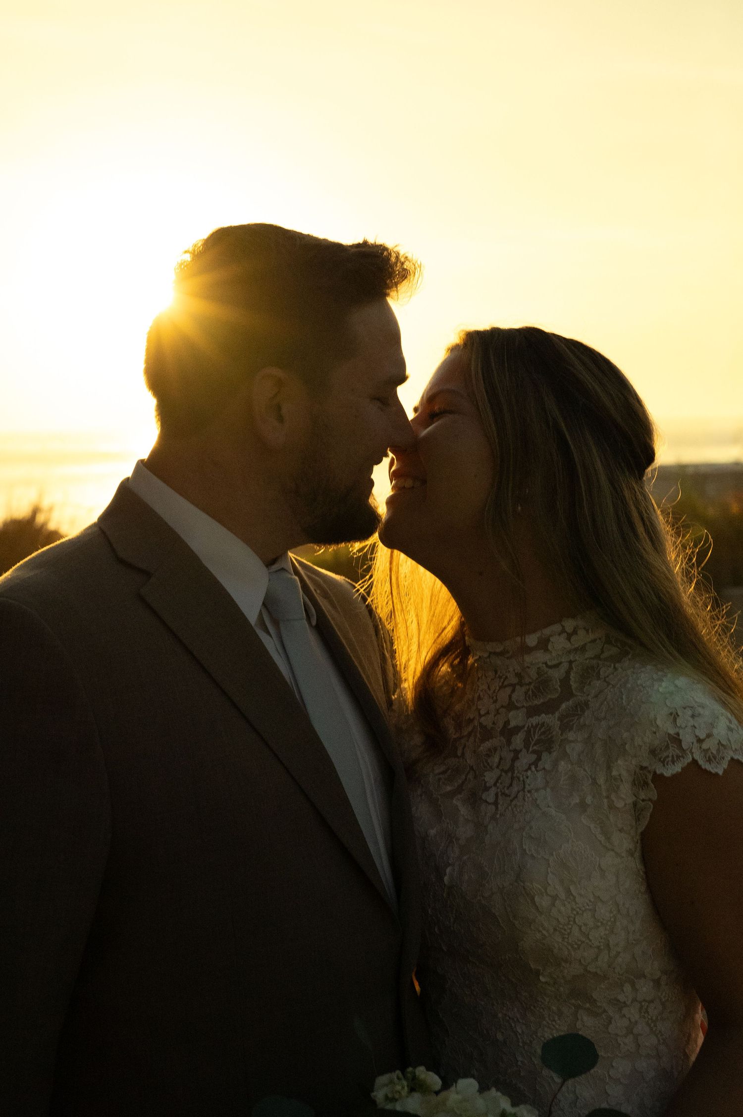 Bride and groom sharing a romantic kiss at golden sunset, backlit by warm glowing light.