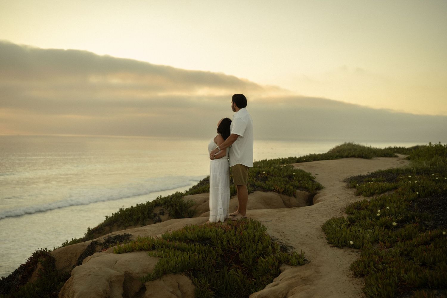 Couple in white attire standing on coastal cliffs watching a serene ocean sunset together.