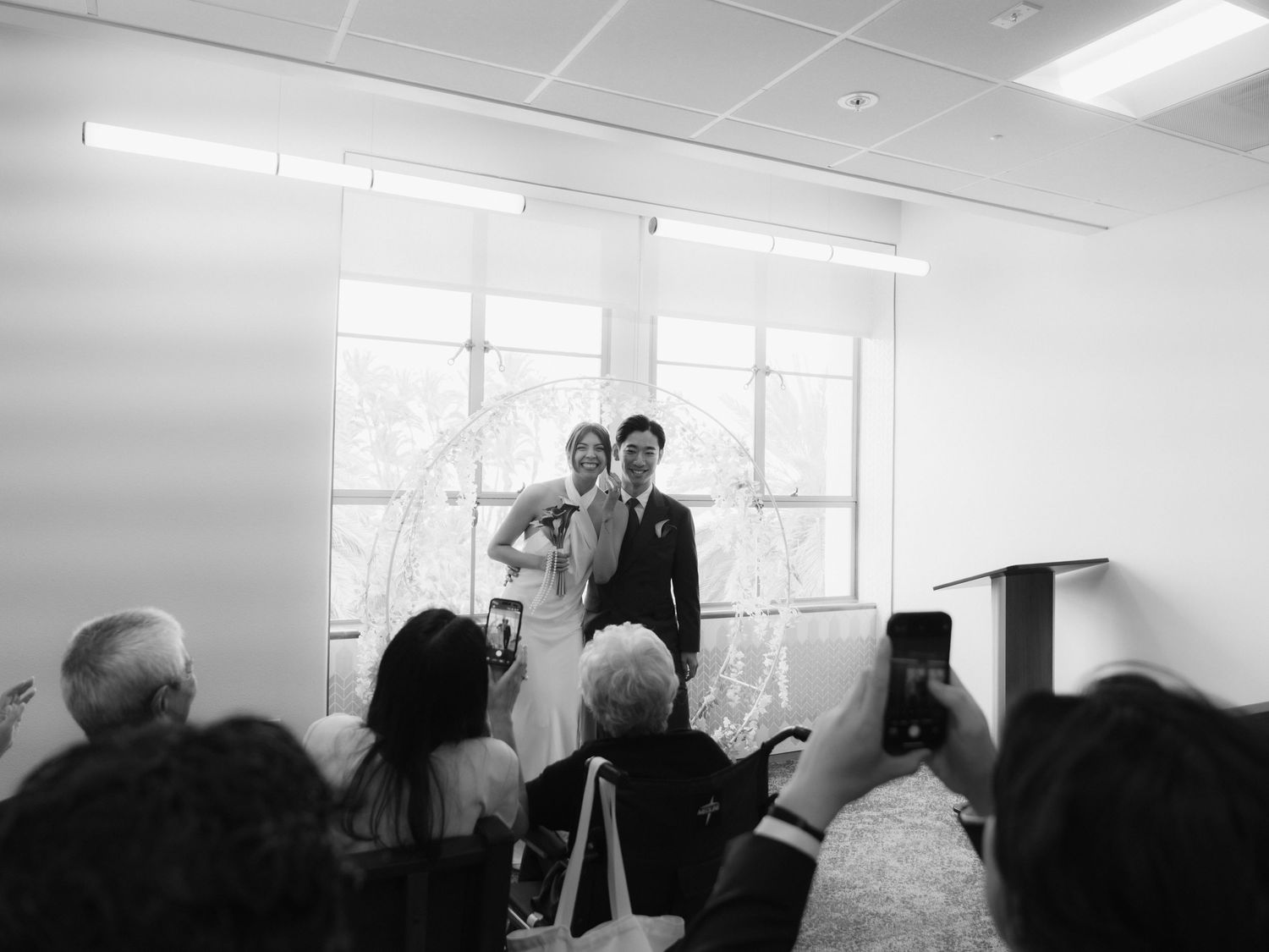 Black and white wedding ceremony photo of bride and groom standing before guests in a bright modern venue.