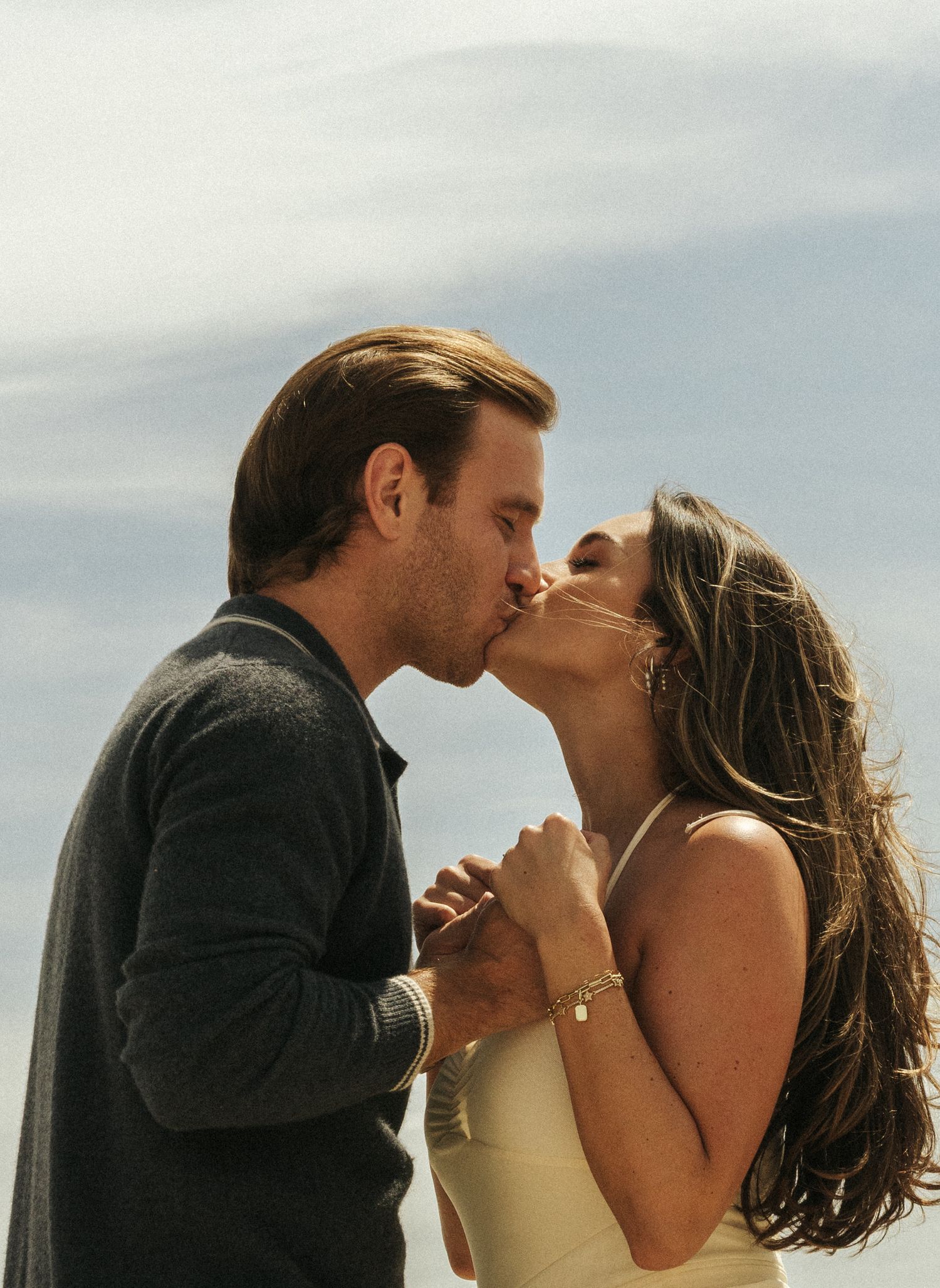 Romantic couple sharing a kiss outdoors while holding hands under a bright cloudy sky.