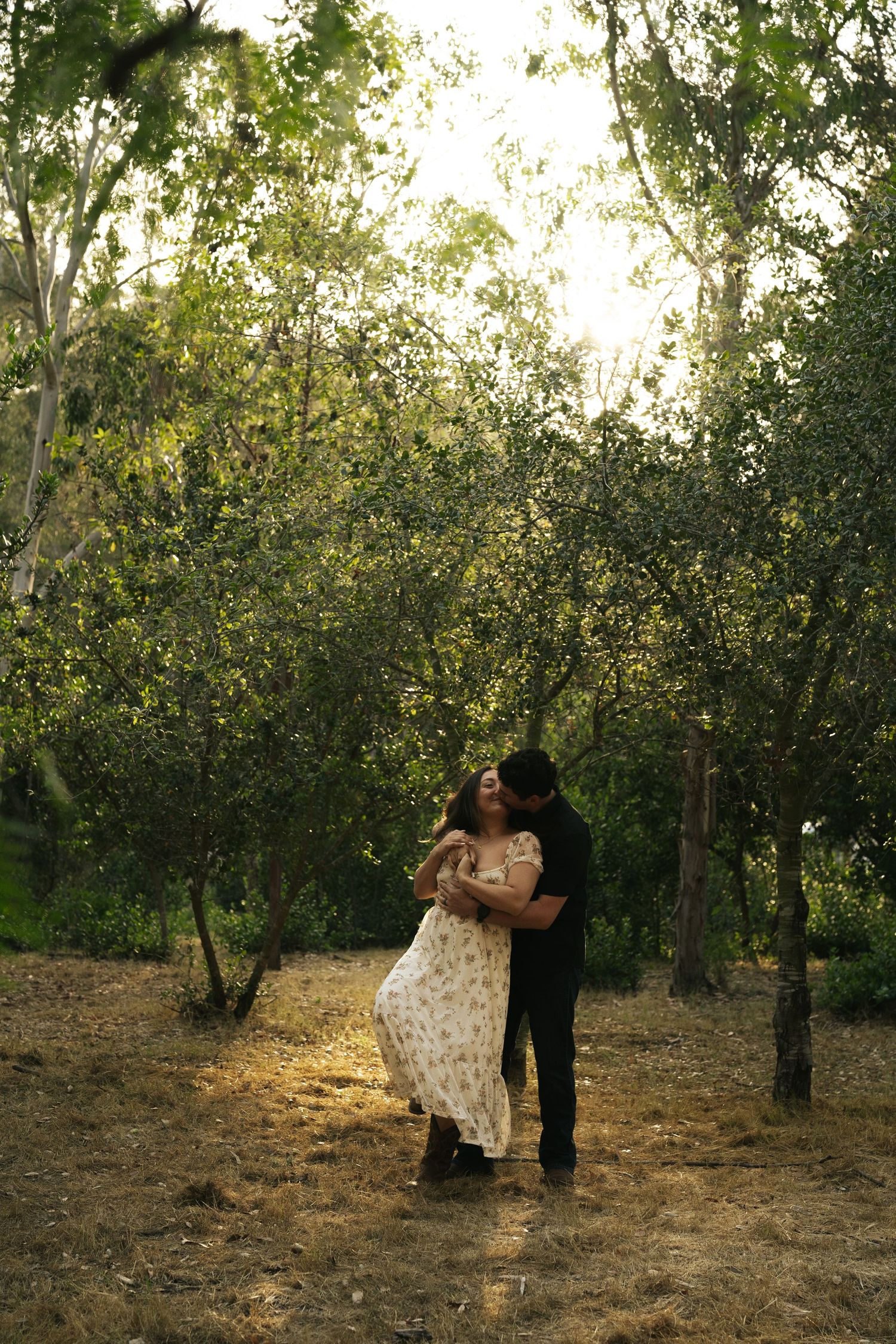 Man in black embracing woman in floral dress in a warmly lit woodland setting at golden hour.