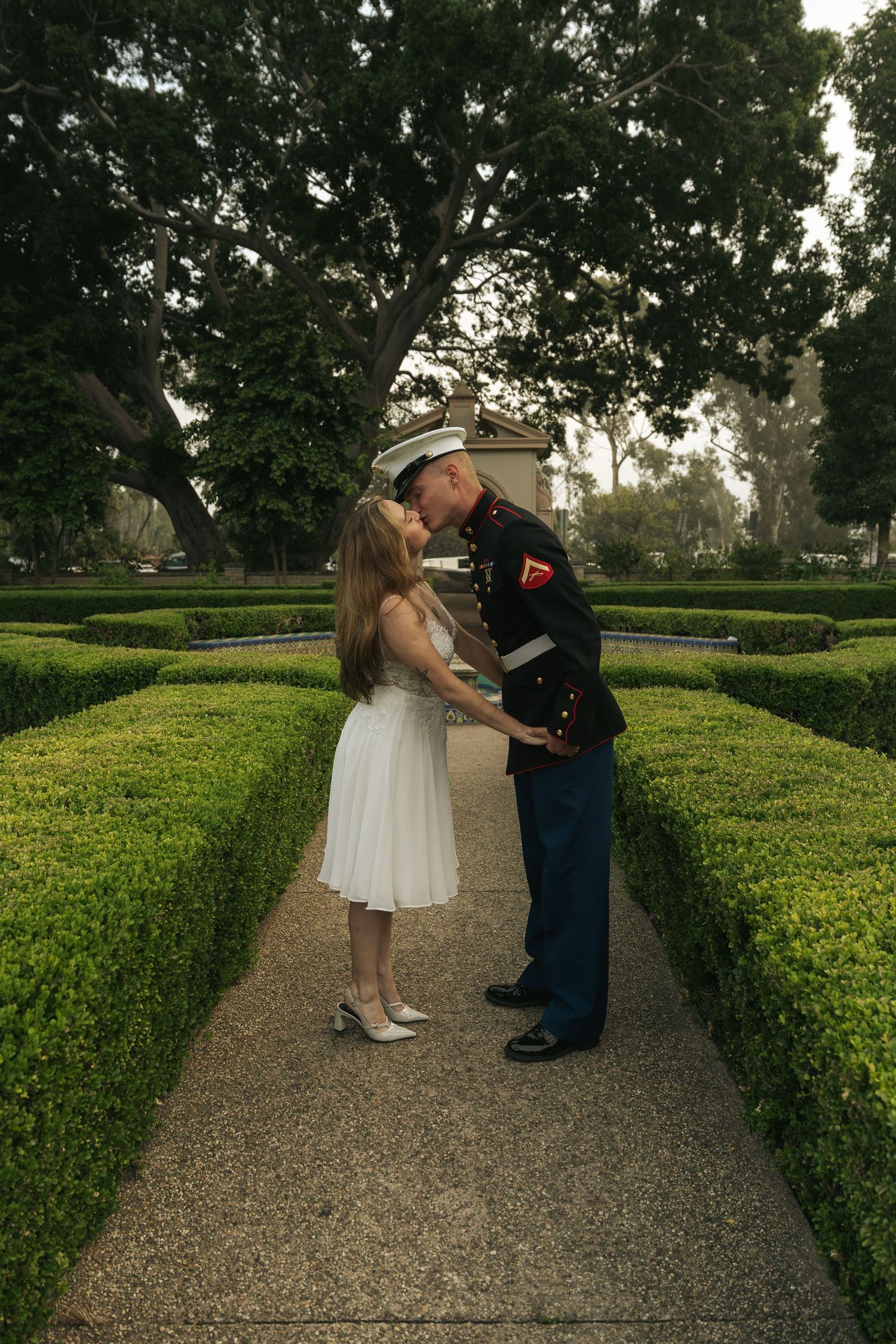 Marine in dress uniform kisses bride in white dress along a manicured hedge garden path.