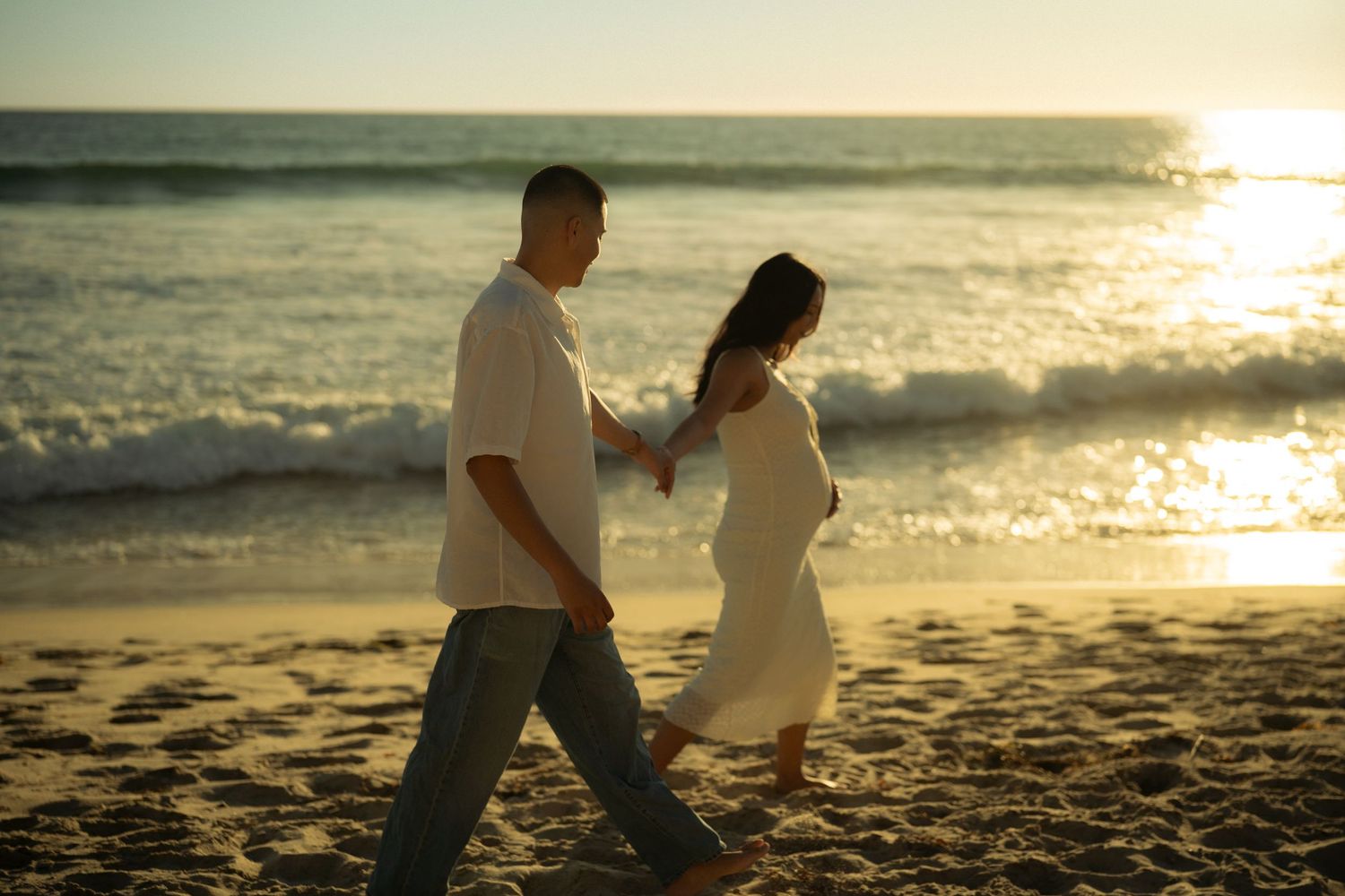 Couple holding hands walking on a sandy beach at golden sunset with ocean waves in the background.