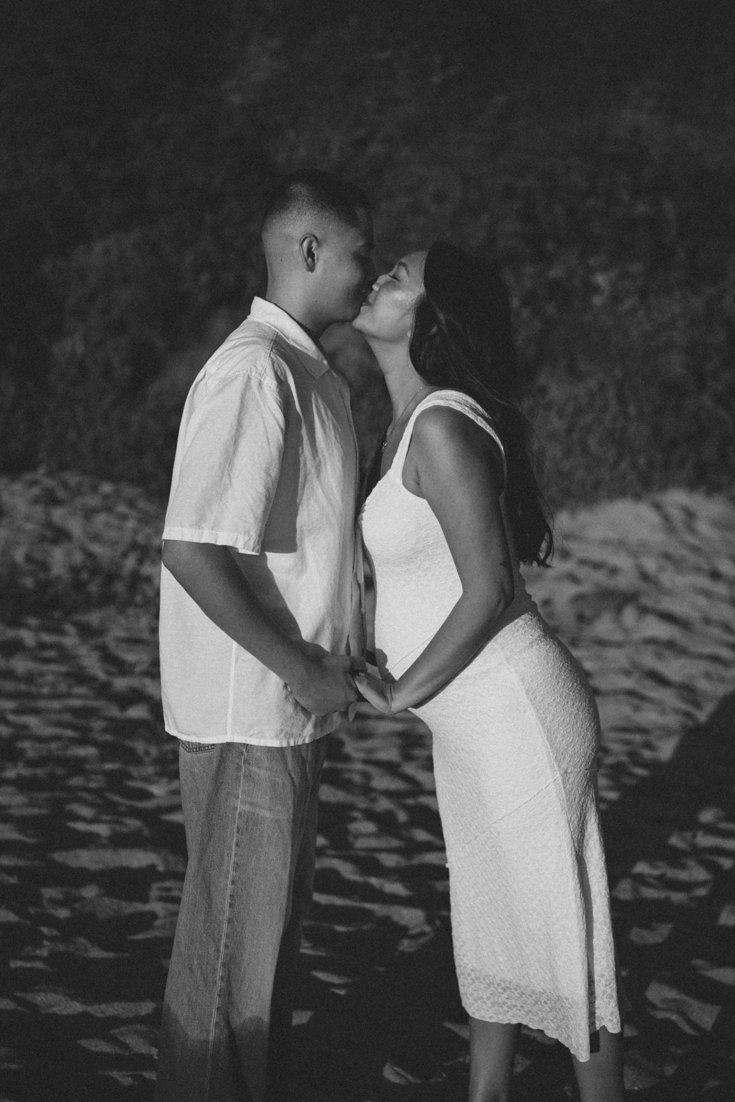 Black and white photo of a couple kissing on a beach while holding hands.