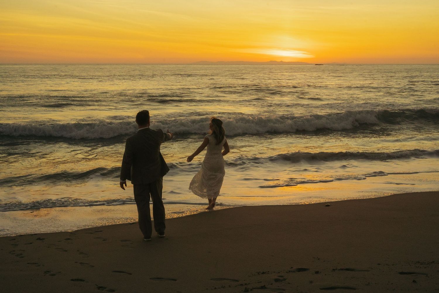 Bride and groom walk hand in hand toward glowing ocean sunset on their wedding day at the beach.