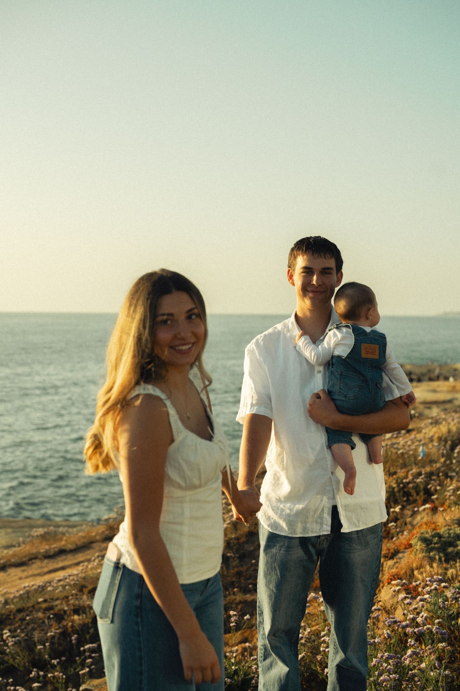 Young family with baby posing by the seaside at sunset, woman in white top and jeans, man holding infant.