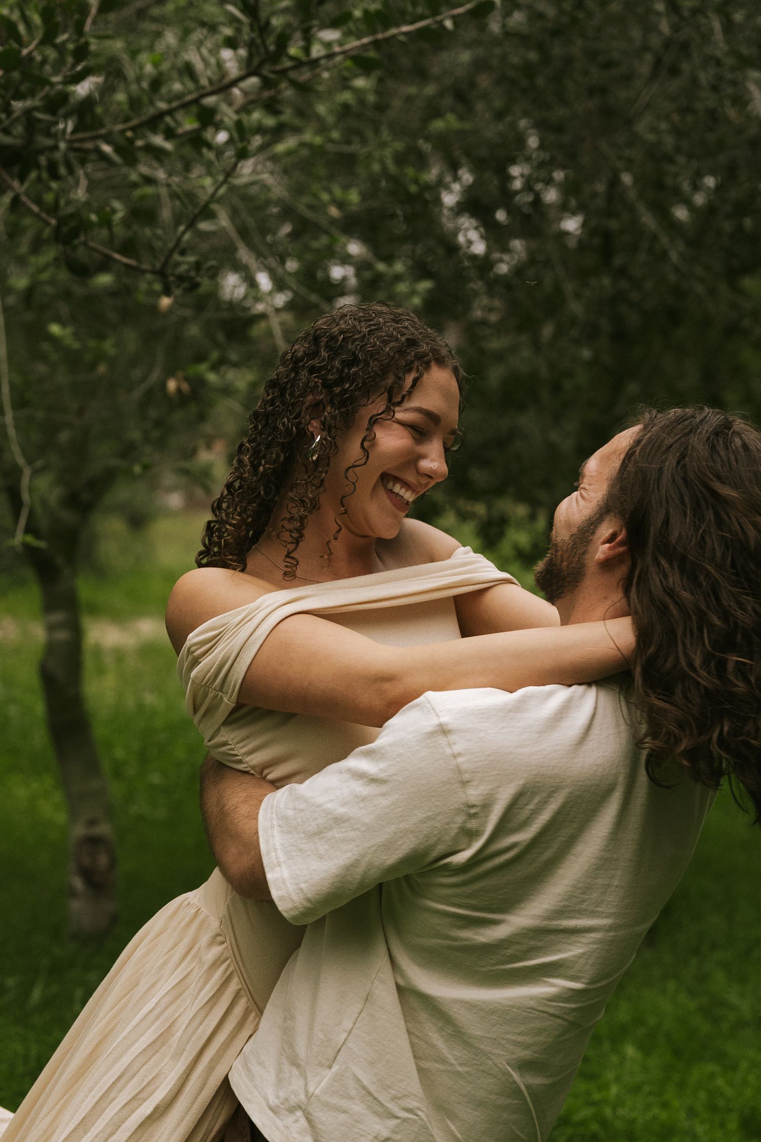 A joyful couple embraces and laughs together in a lush green garden, wearing elegant cream-colored outfits.